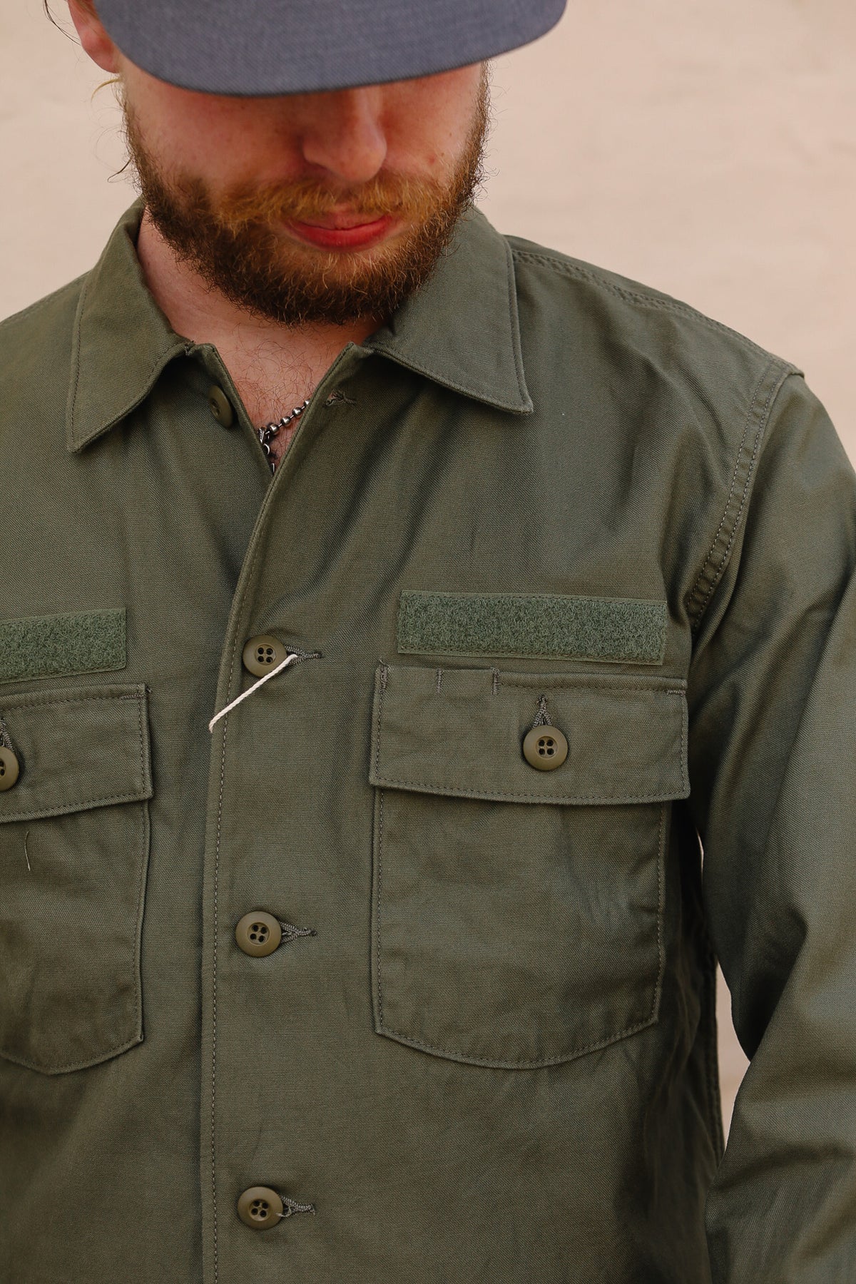 Close-up of a man wearing an olive green military-style button-up shirt jacket with chest pockets and a gray cap, looking down.