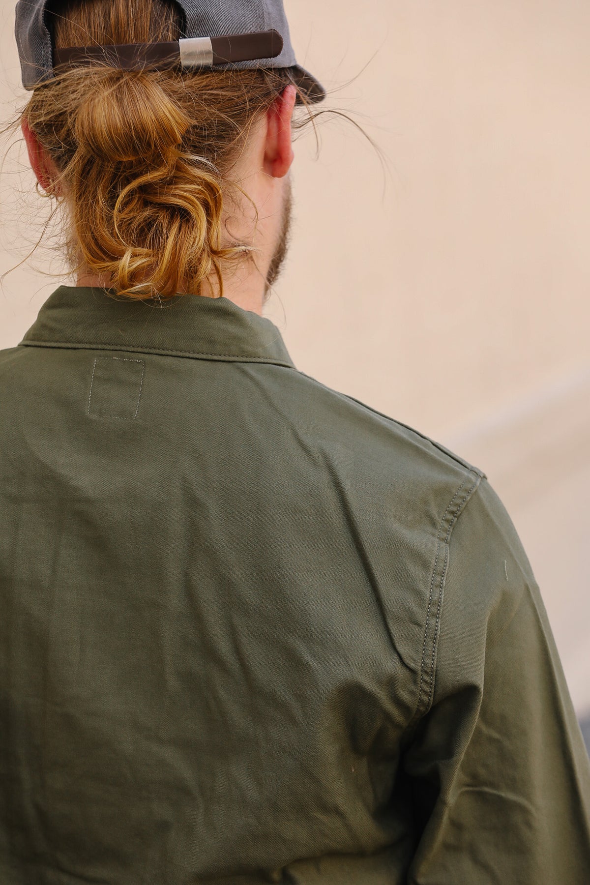 A man viewed from behind wearing an olive green button-up shirt and a dark gray baseball cap, with his hair tied back in a low bun.