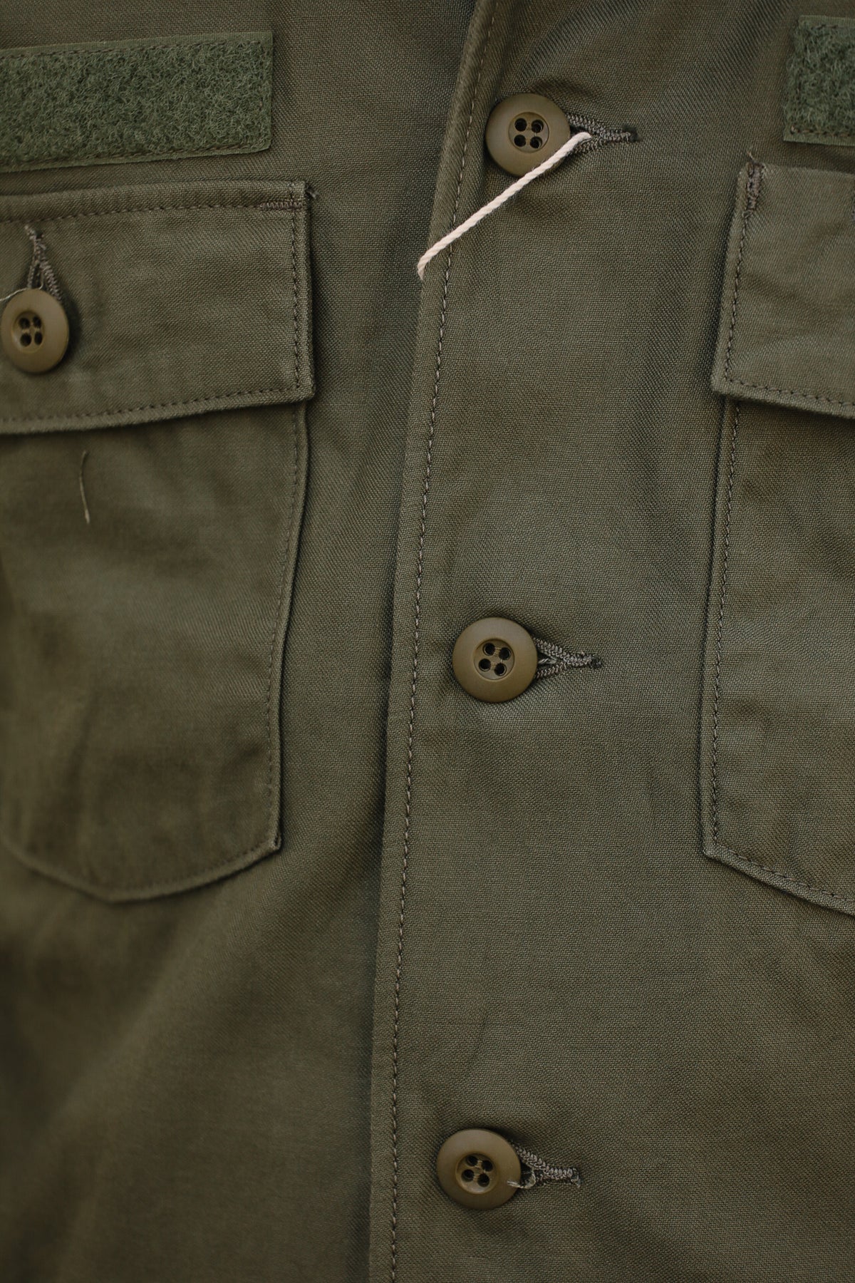 Close-up of an olive green military-style jacket featuring four brown buttons, flap pockets, and a green hook-and-loop patch on the chest.