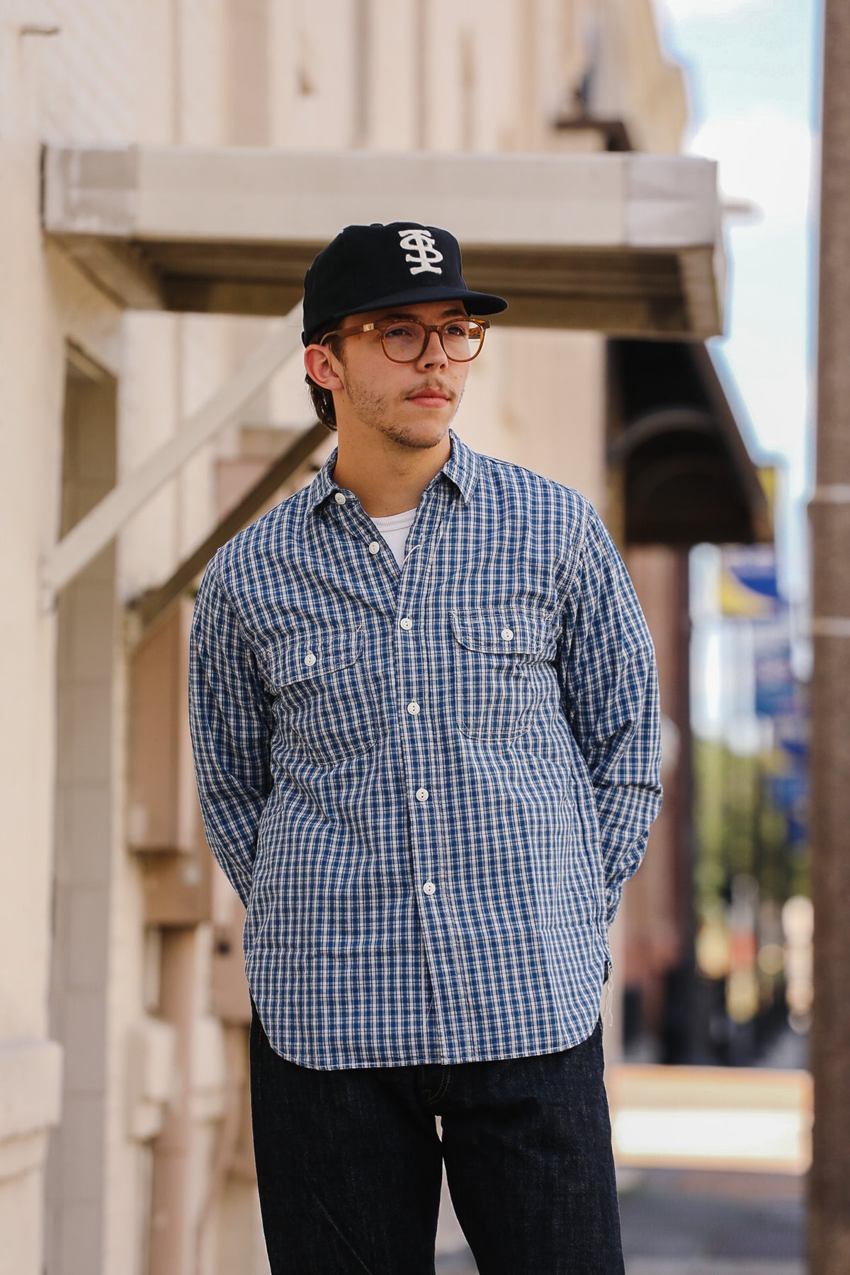 A man wearing a blue and white gingham long-sleeve button-down shirt, dark jeans, a black baseball cap with a white logo, and glasses, standing outdoors in front of a building.