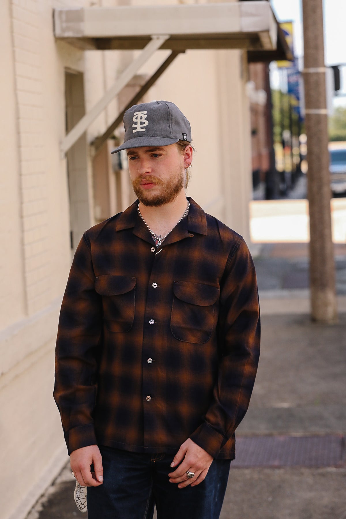 A man wearing a dark brown and black plaid button-up shirt, a gray baseball cap with a white logo, and dark jeans, standing outdoors on a sidewalk next to a beige brick building.