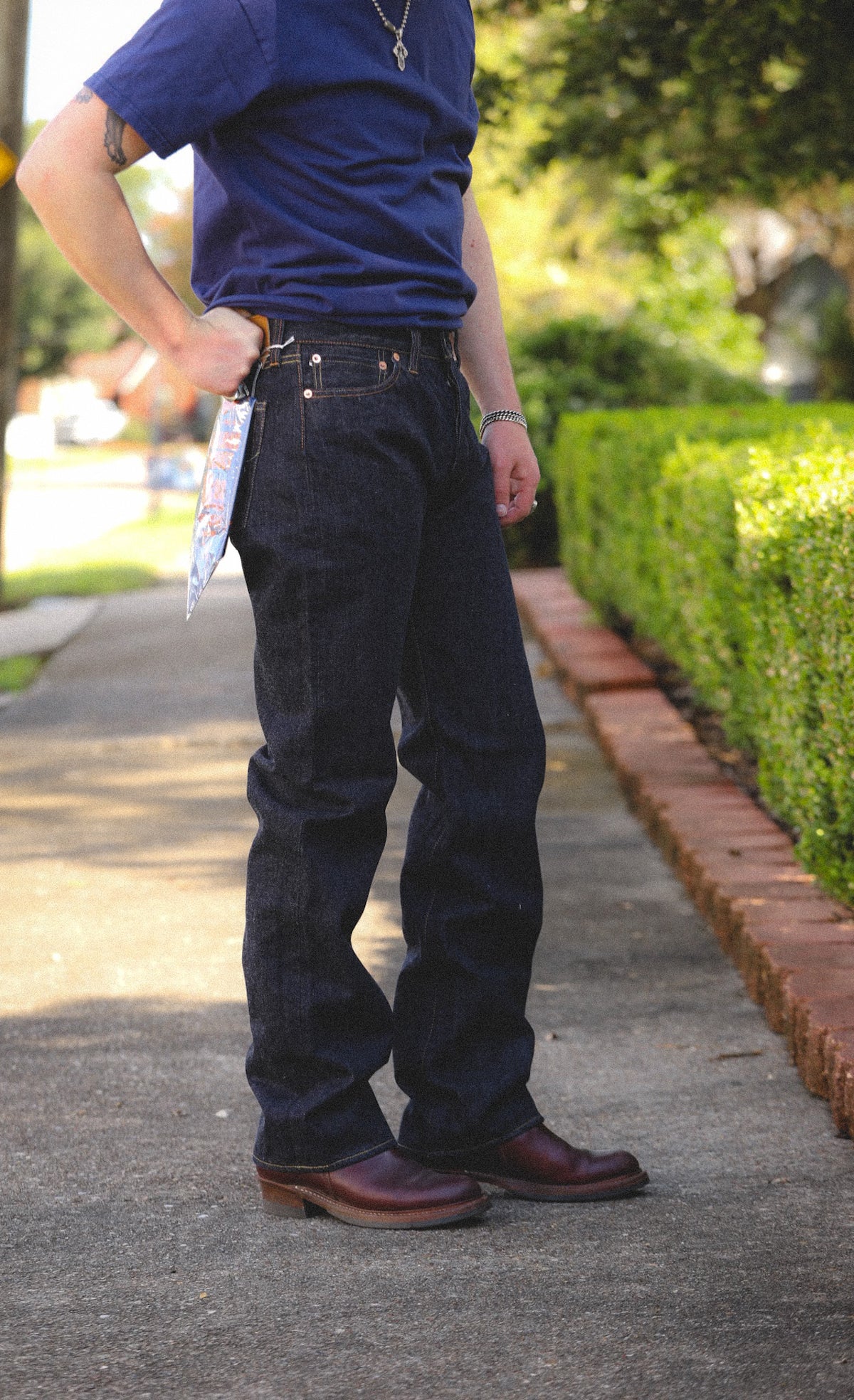 A person wearing dark indigo jeans, a navy blue t-shirt, and brown leather boots, standing on a paved path outdoors next to a green hedge.