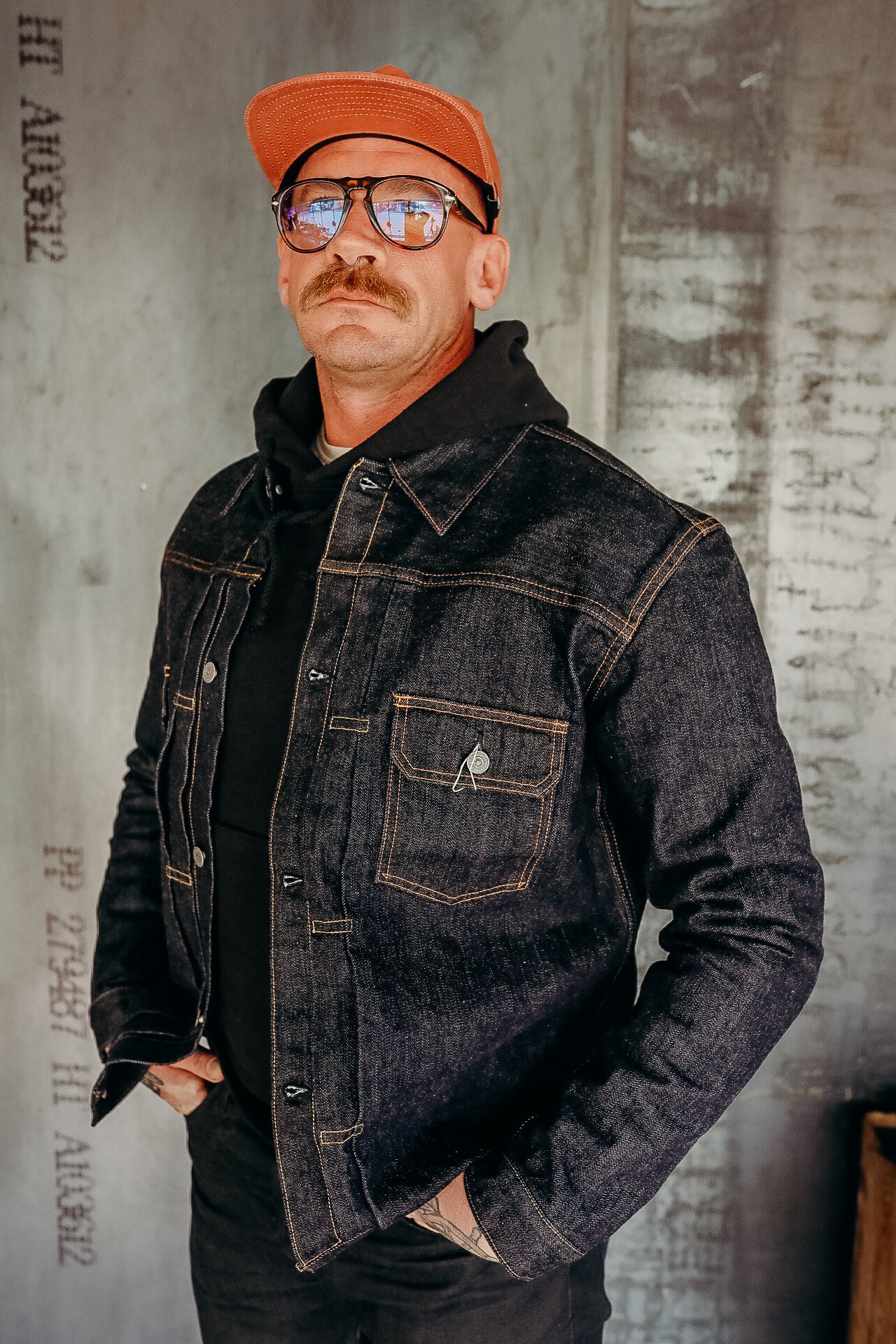 Man with mustache wearing an orange cap, sunglasses, black hoodie, and dark denim jacket against concrete wall