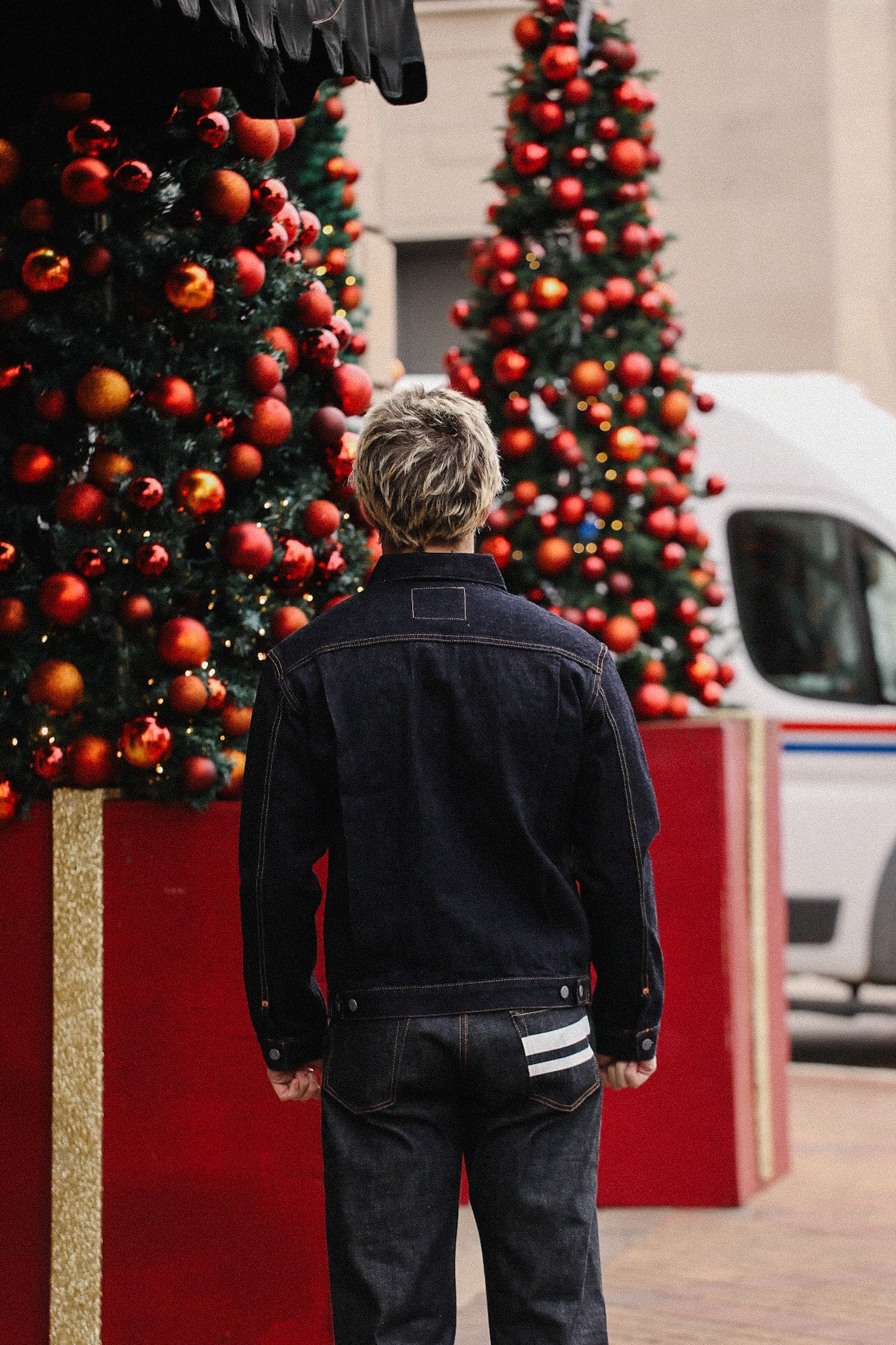 Man in black denim jacket and jeans standing near festive Christmas trees decorated with red ornaments