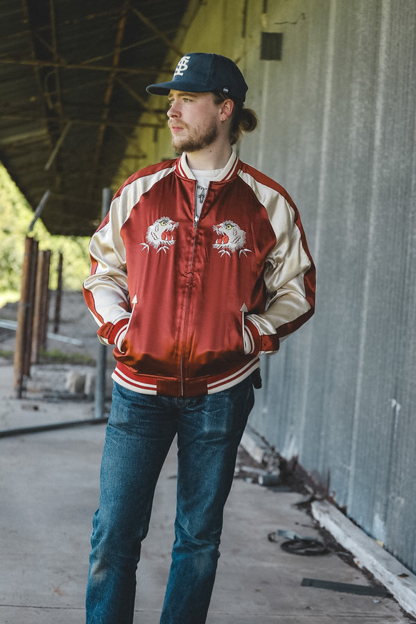 A man wearing a red and beige varsity bomber jacket with embroidered eagle designs on the chest, blue jeans, and a baseball cap, standing in an industrial setting.