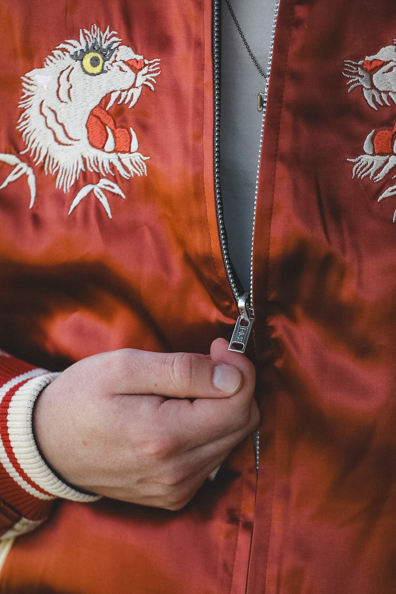 Close-up of a person wearing a shiny rust-orange satin jacket with embroidered tiger faces and pulling a silver zipper with one hand.