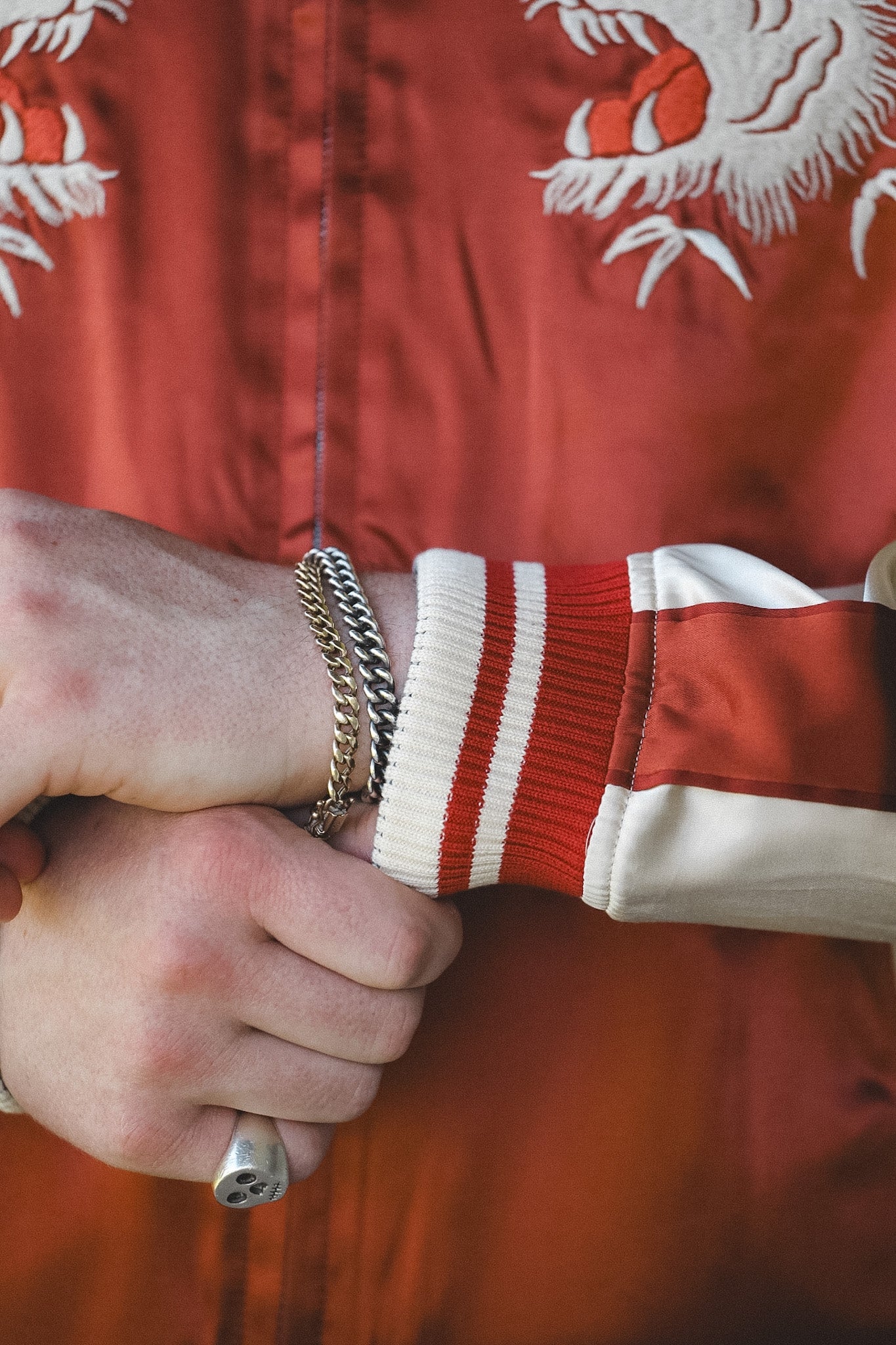 Close-up of a person wearing a red satin bomber jacket with white embroidered dragon motifs and red-and-white striped ribbed cuffs, while adjusting the collar. A silver chain bracelet and ring are visible on the wrist.