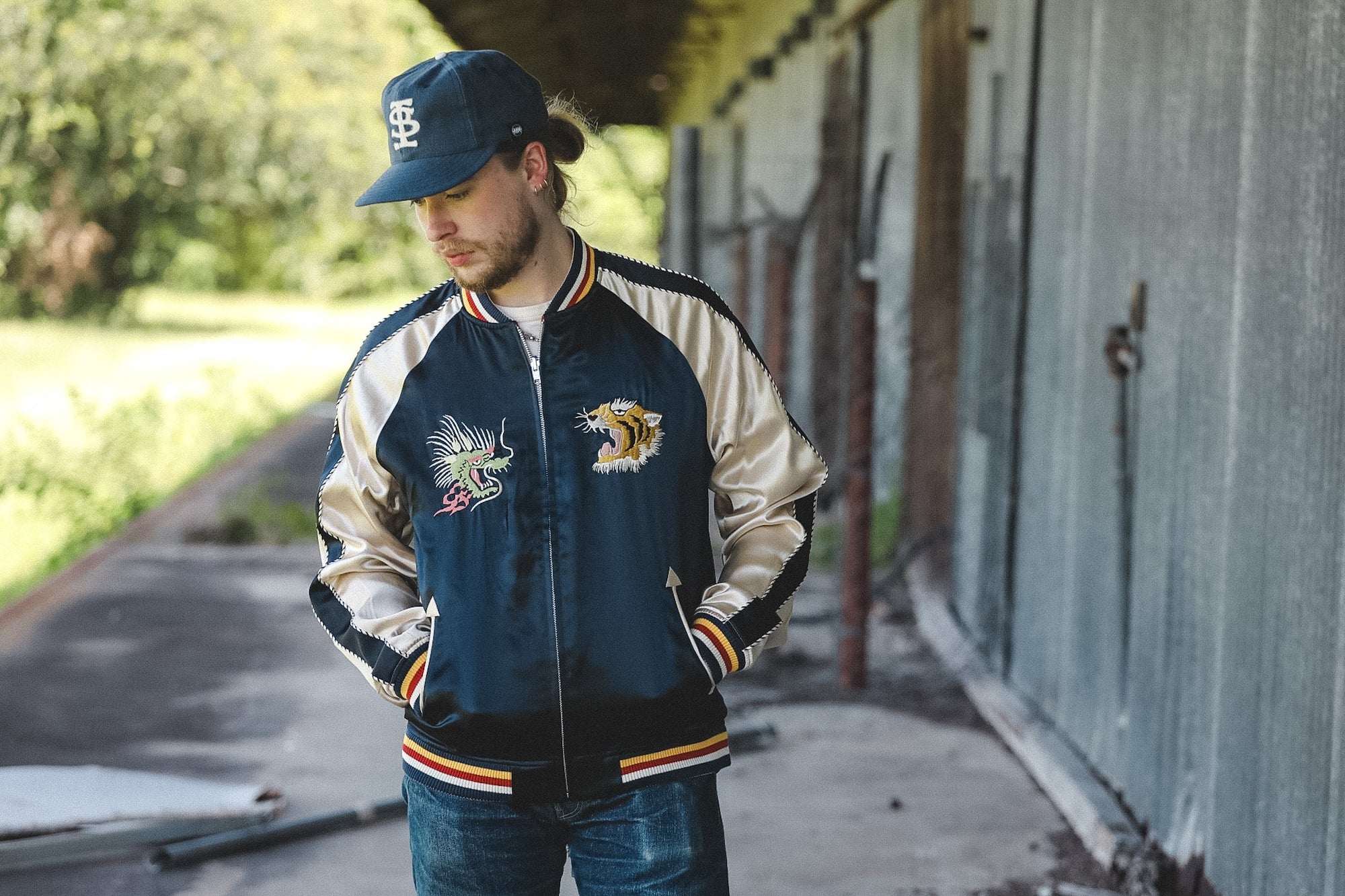 A man wearing a navy and beige varsity-style bomber jacket with embroidered patches, a matching navy baseball cap, and blue jeans, standing outdoors next to a weathered metal building.
