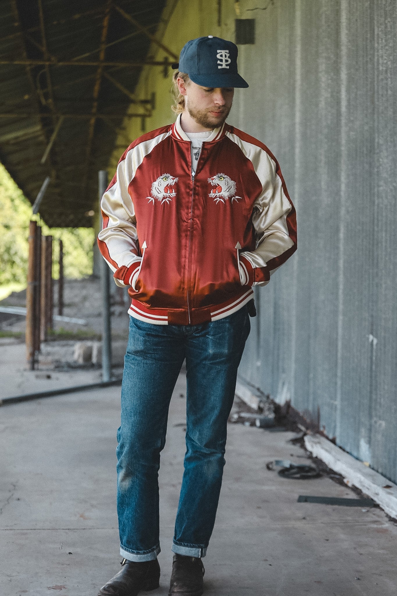 A man wearing a red and cream satin bomber jacket with embroidered dragon designs on the chest, blue jeans, and a navy baseball cap, standing in an industrial setting.