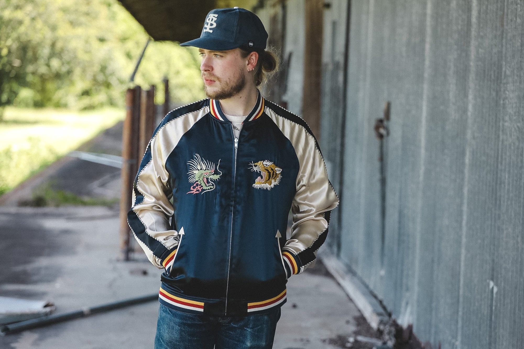 A man wearing a navy and beige varsity-style bomber jacket with embroidered graphics, a matching navy baseball cap, and blue jeans, standing outdoors against a gray metal building.