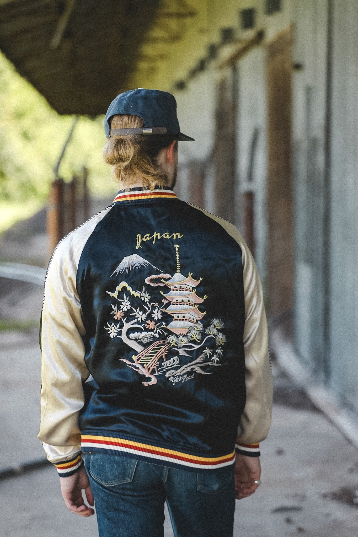 A man viewed from behind wearing a navy and beige satin bomber jacket with an intricate Japanese pagoda and Mount Fuji embroidery on the back, walking along an outdoor path.