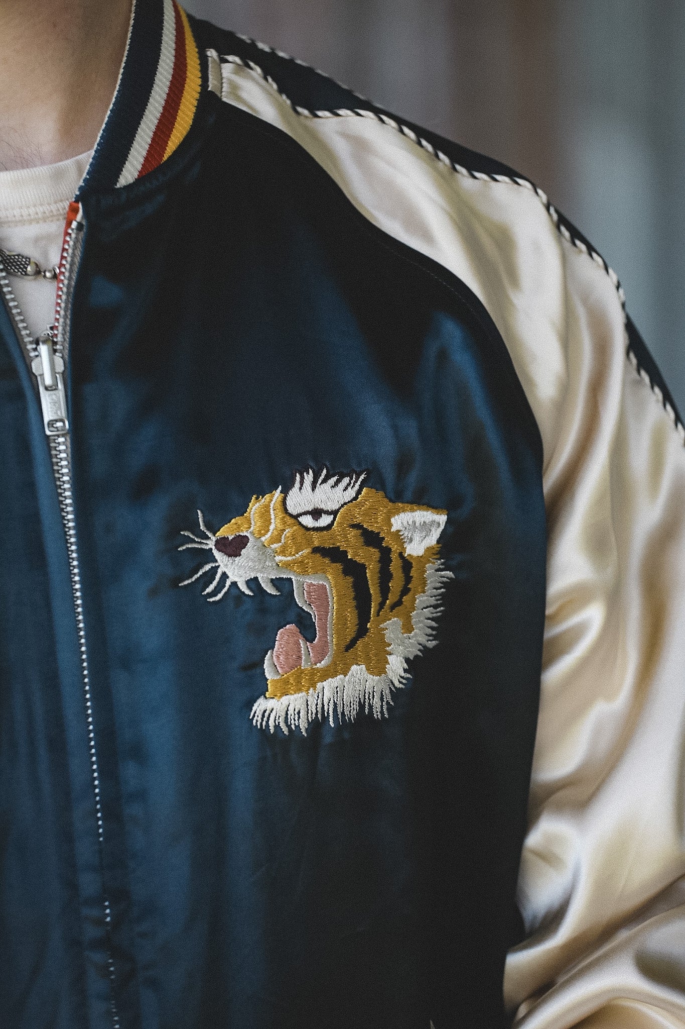 Close-up of a navy satin bomber jacket with cream satin sleeves, a striped ribbed collar, and an embroidered roaring tiger face on the chest.