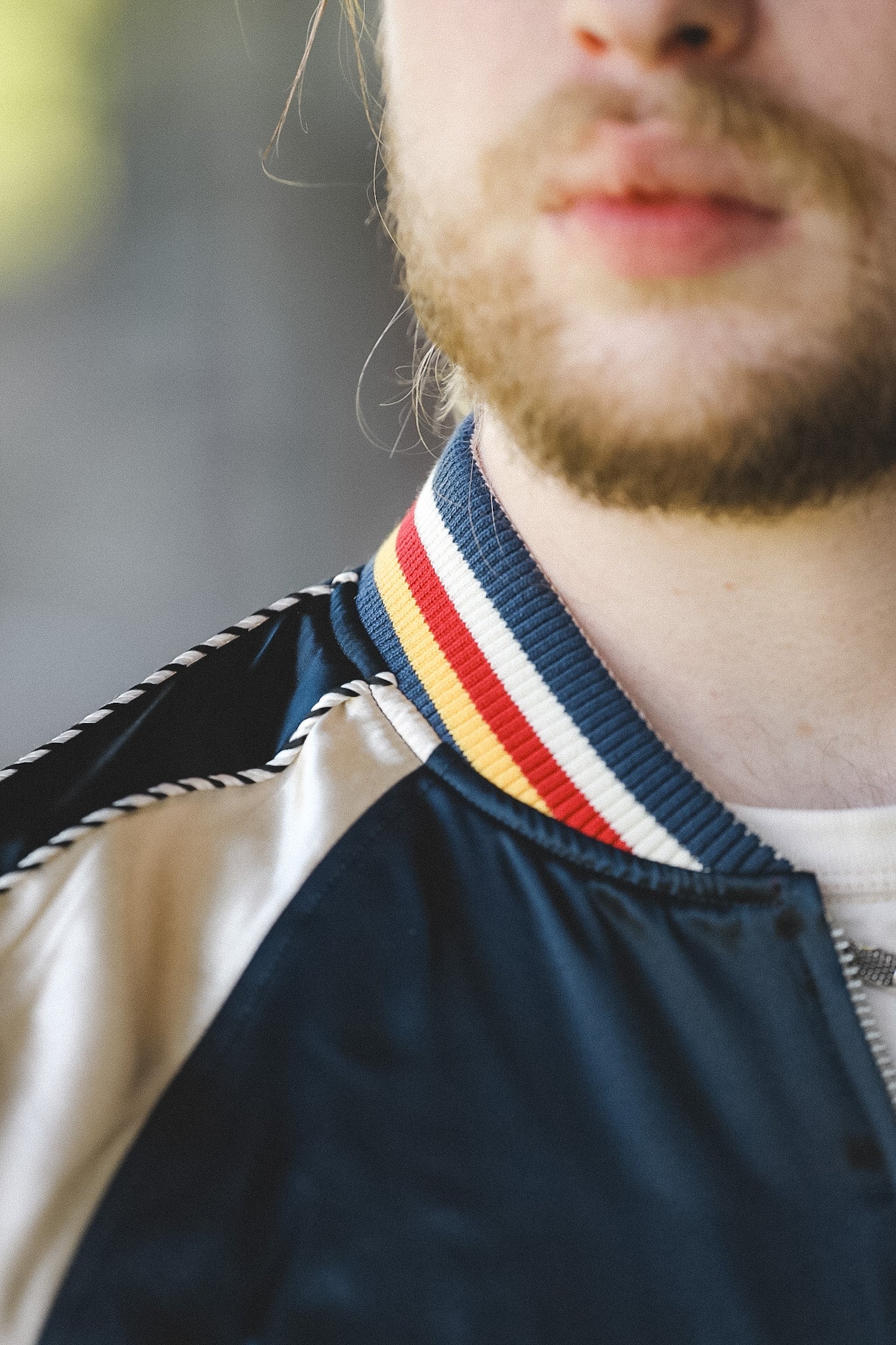 Close-up of a person wearing a navy satin bomber jacket with cream-colored satin shoulder panels and a striped ribbed collar in navy, white, yellow, and red.