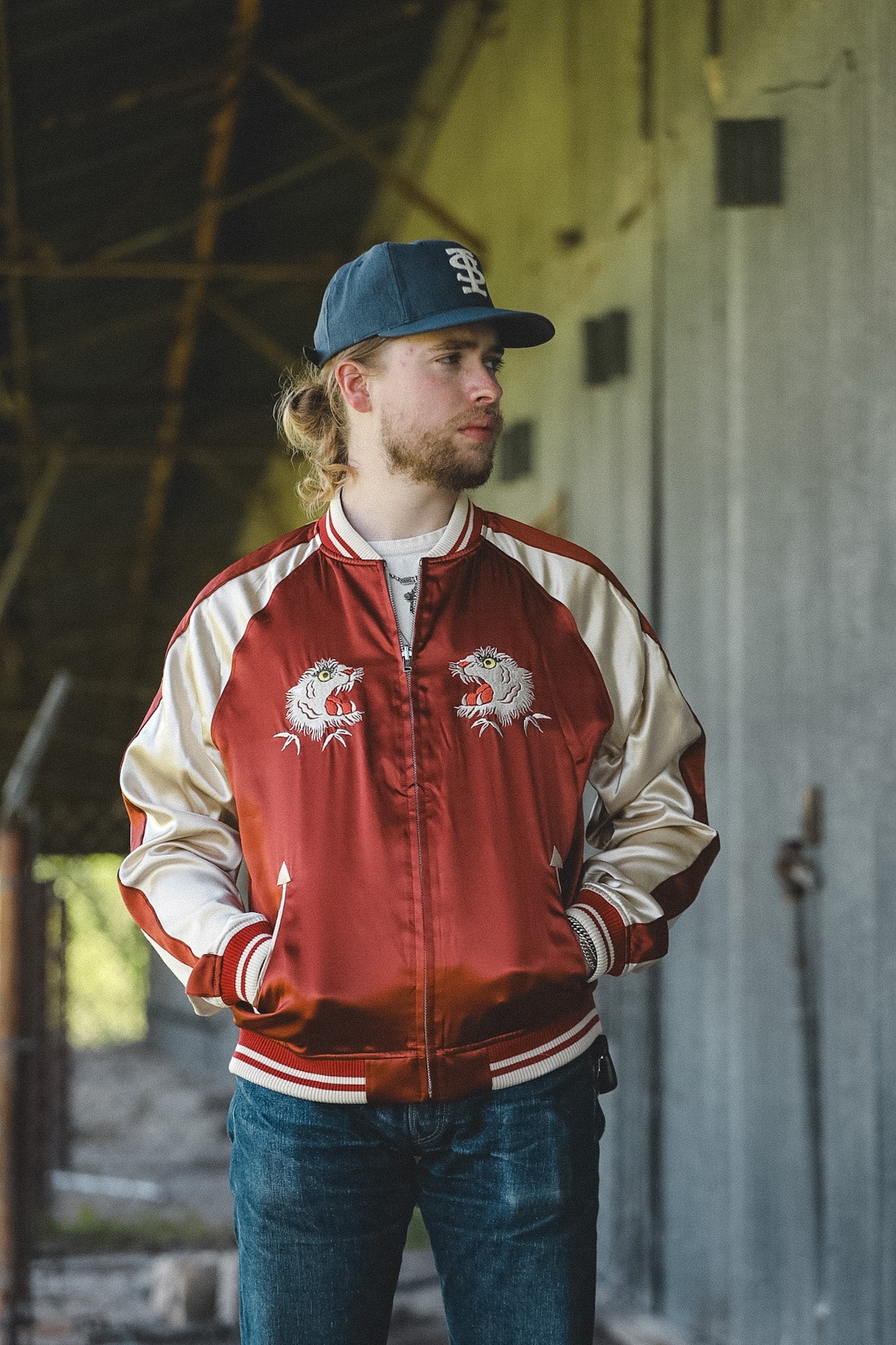 A man wearing a red and cream satin bomber jacket with embroidered tiger designs on the chest, blue jeans, and a blue baseball cap, standing in a rustic indoor setting.