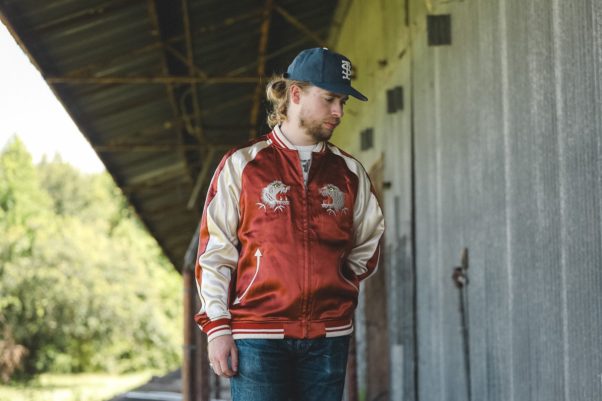 A man wearing a red and beige satin bomber jacket with embroidered eagle designs, blue jeans, and a dark blue baseball cap, standing under a rustic metal roof next to a corrugated metal wall.