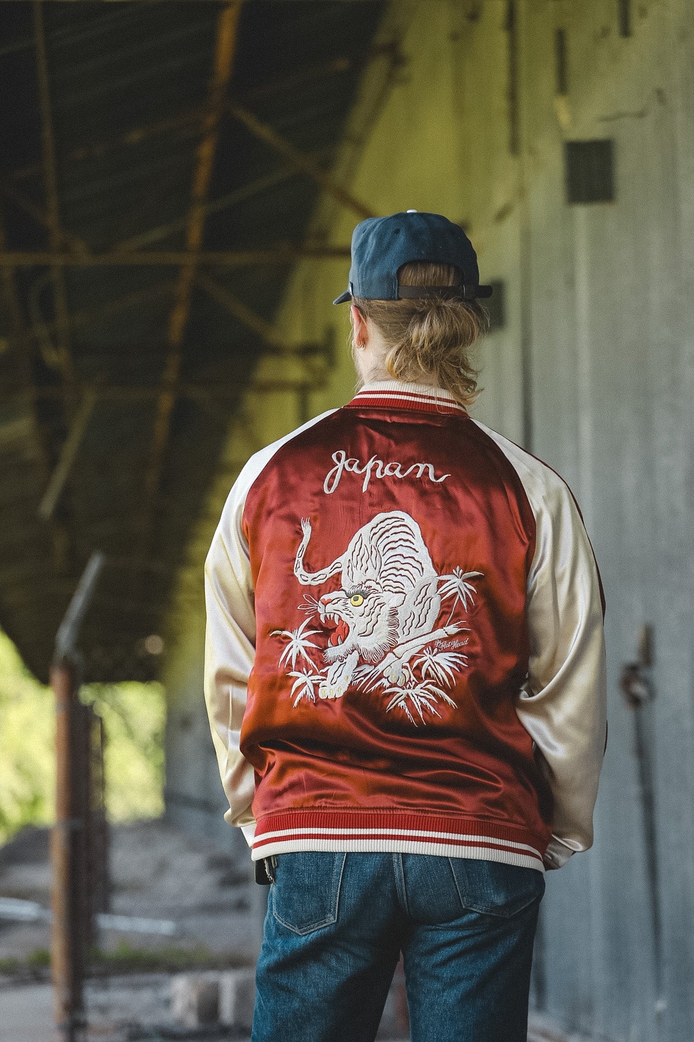 A person viewed from behind wearing a red and cream varsity-style jacket with a large embroidered tiger design and the word 'Japan' on the back, standing in an industrial interior.