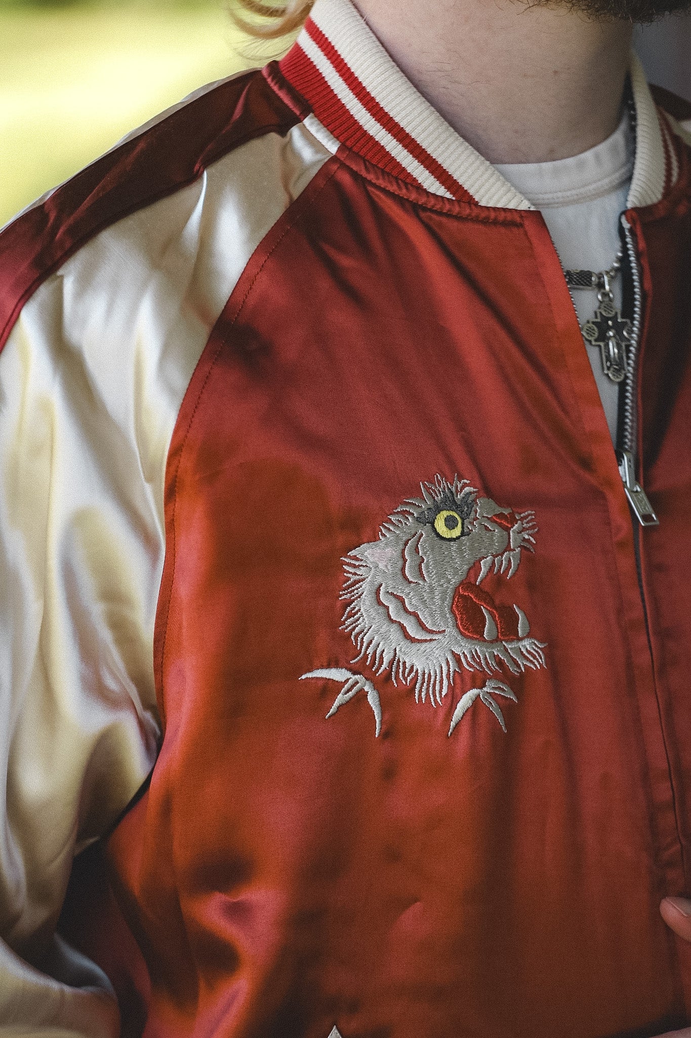 A close-up of a person wearing a satin bomber jacket with cream-colored sleeves and collar, featuring a red body and a white embroidered monster design on the chest.