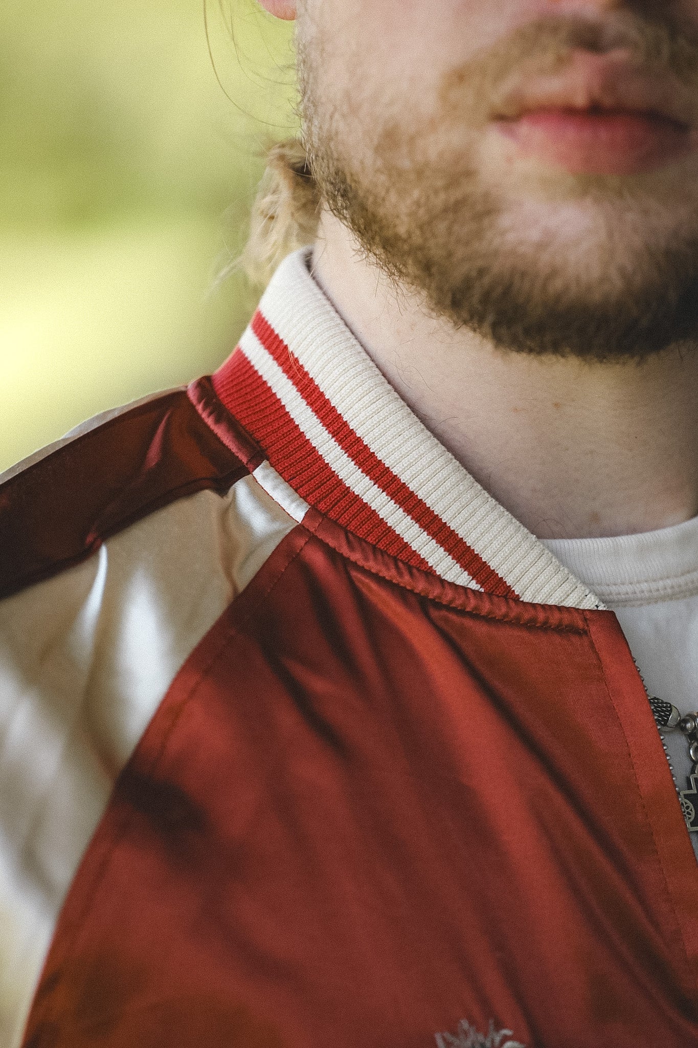 Close-up of a person wearing a satin bomber jacket with a red and white striped ribbed collar, a white crewneck shirt underneath, and a blurred green outdoor background.