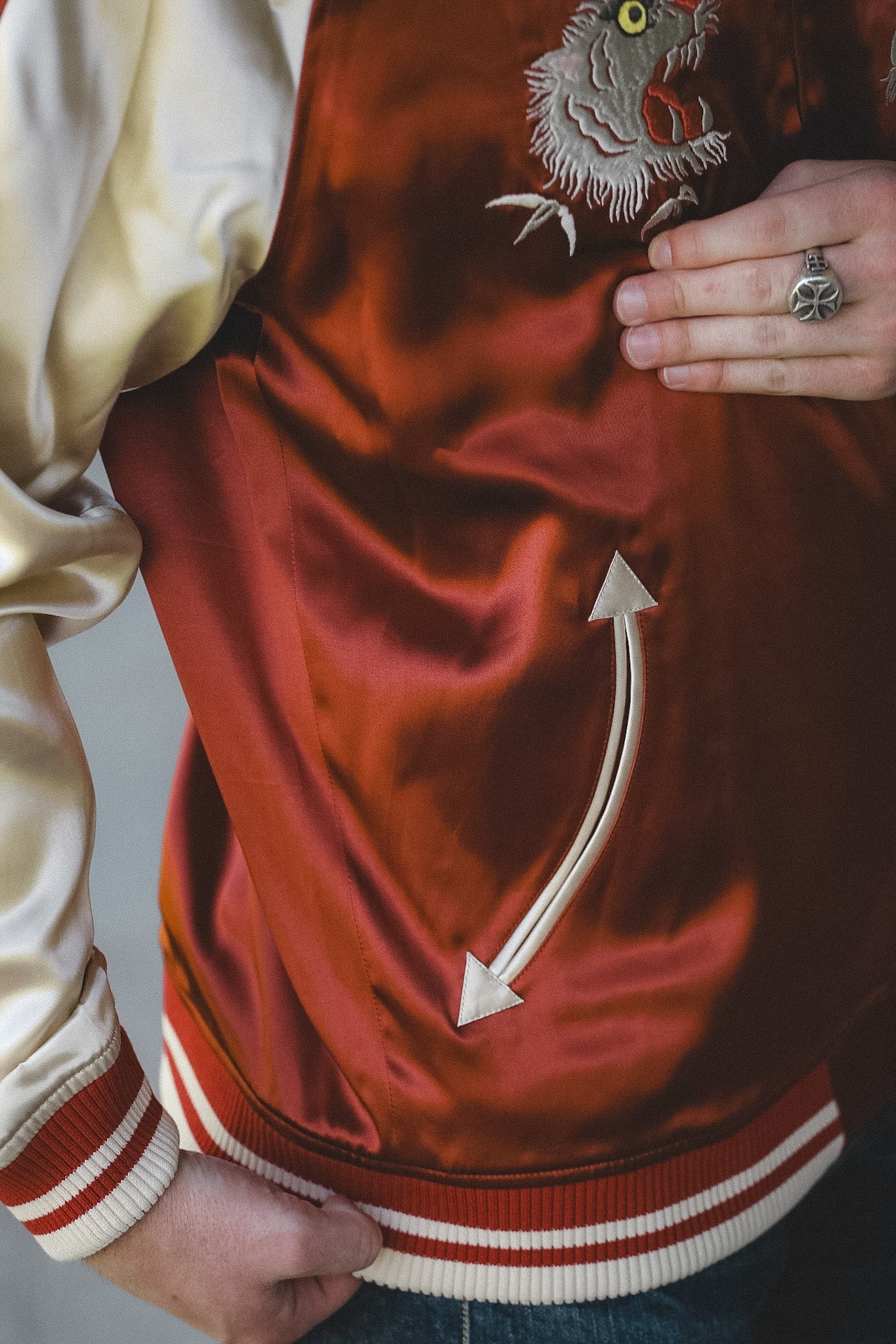 Close-up of a satin varsity jacket worn by a person, featuring a rust red body with cream ribbed trim, cream satin sleeves, and a large embroidered owl on the chest.