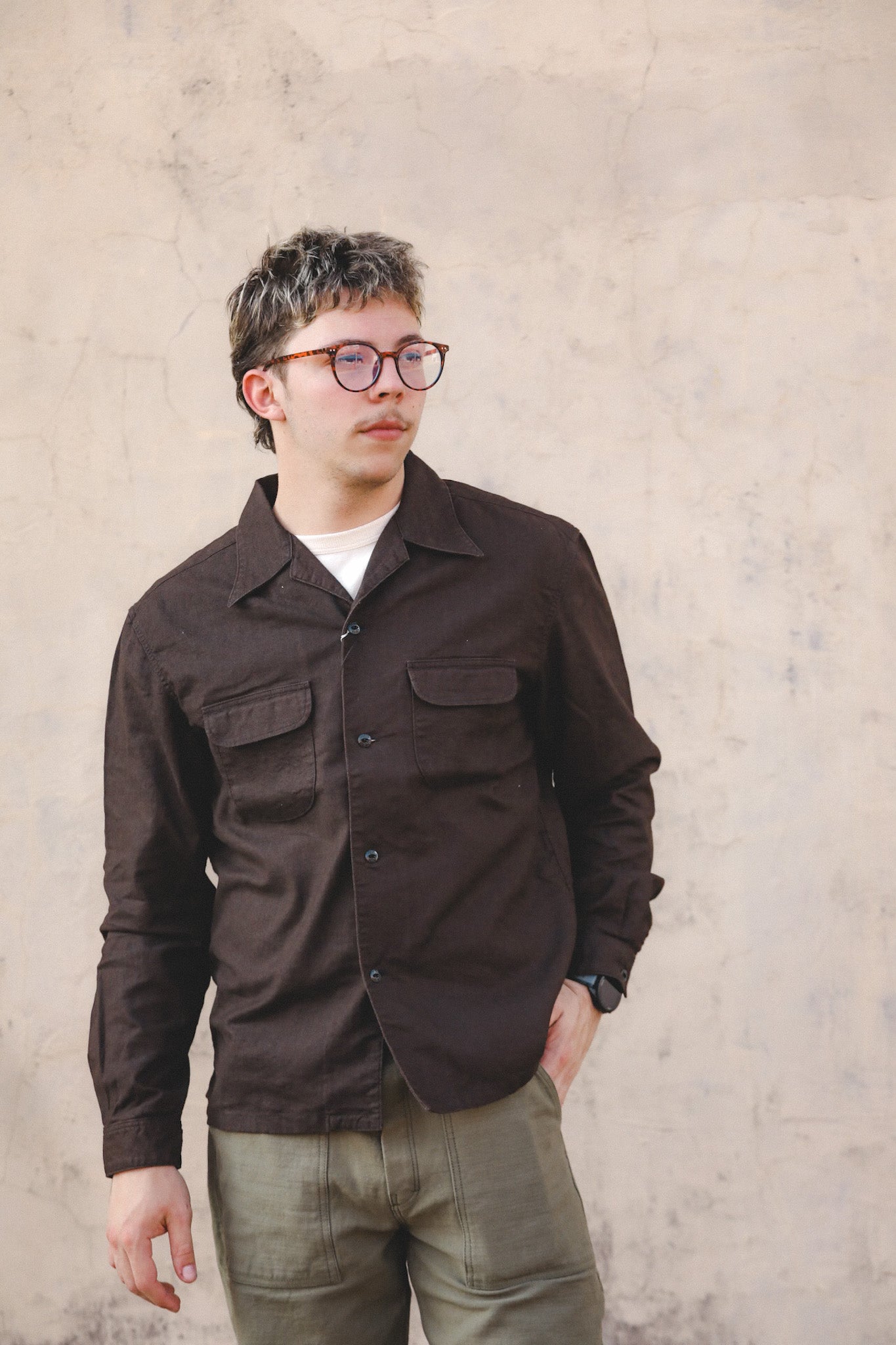 Man wearing a brown jacket and glasses against a beige wall