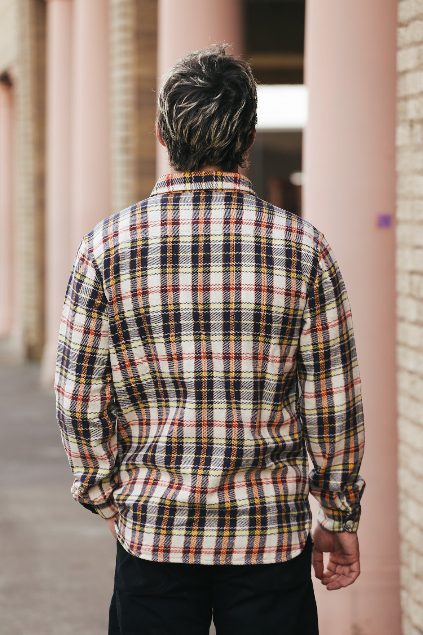 Person wearing a plaid shirt standing in front of architectural columns.