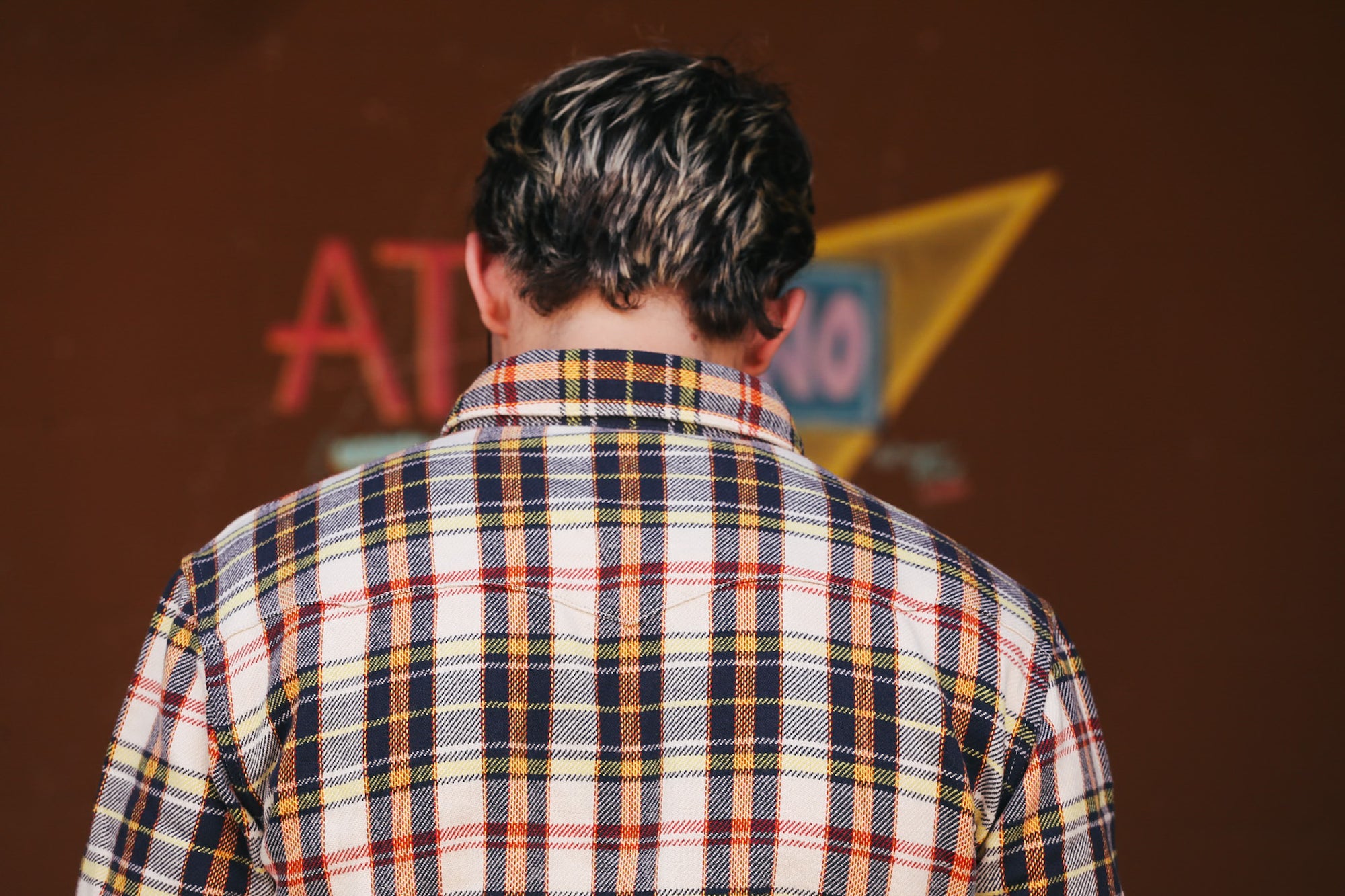 Person wearing a plaid shirt with a blurred background featuring colorful signs.