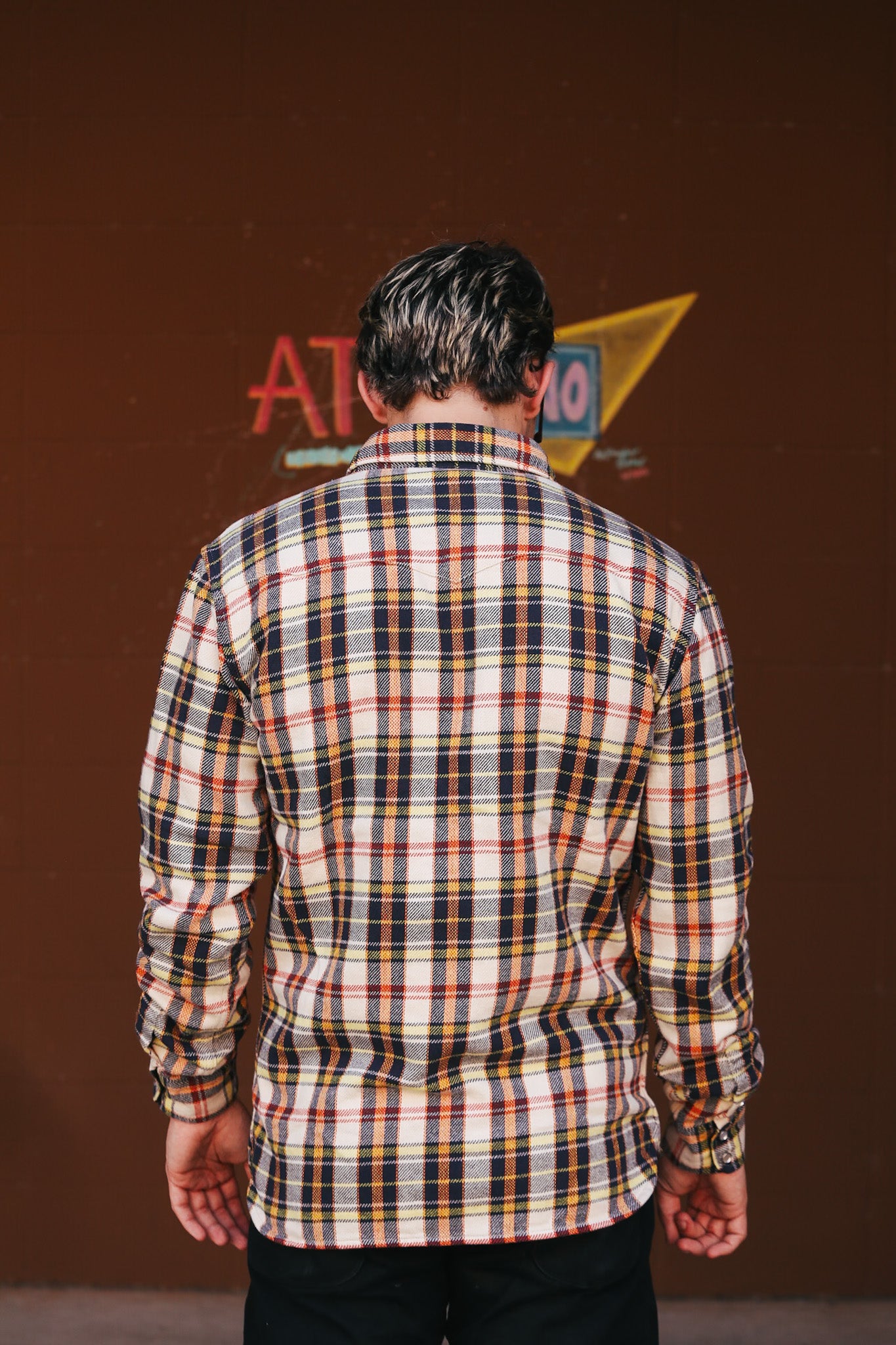 Person wearing a plaid shirt with a brown wall and vintage sign in the background