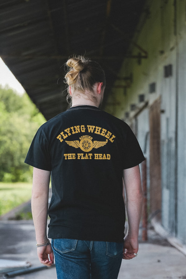 A person viewed from behind wearing a black t-shirt with a yellow graphic that reads 'FLYING WHEEL' and 'THE FLAT HEAD' with an airplane wing emblem. The person is walking along an outdoor covered walkway next to a building.