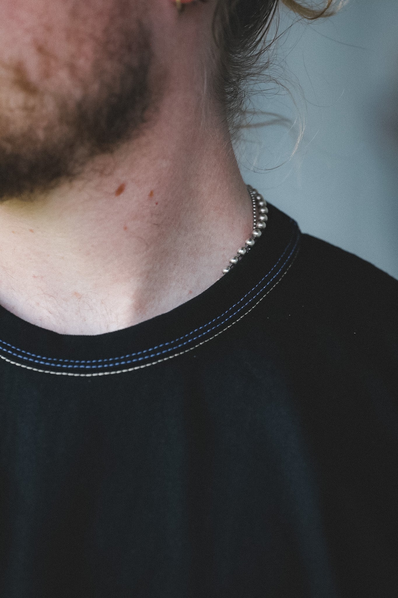 Close-up of a person's neck and upper chest wearing a thin silver beaded necklace and a black crew-neck t-shirt with blue contrast stitching.