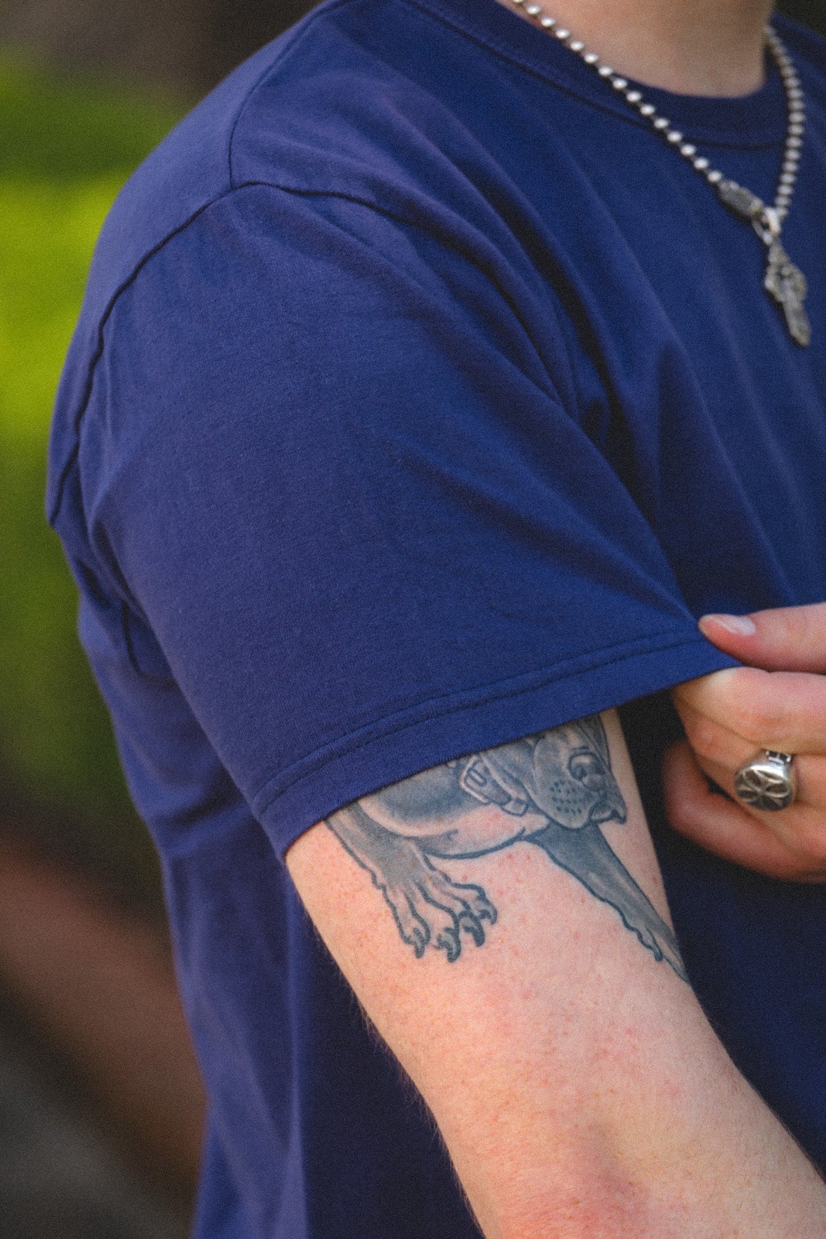 A close-up of a person wearing a navy blue t-shirt, showing a detailed black and gray tattoo of a dog on their upper arm. The person is also wearing a silver chain necklace and a silver ring.