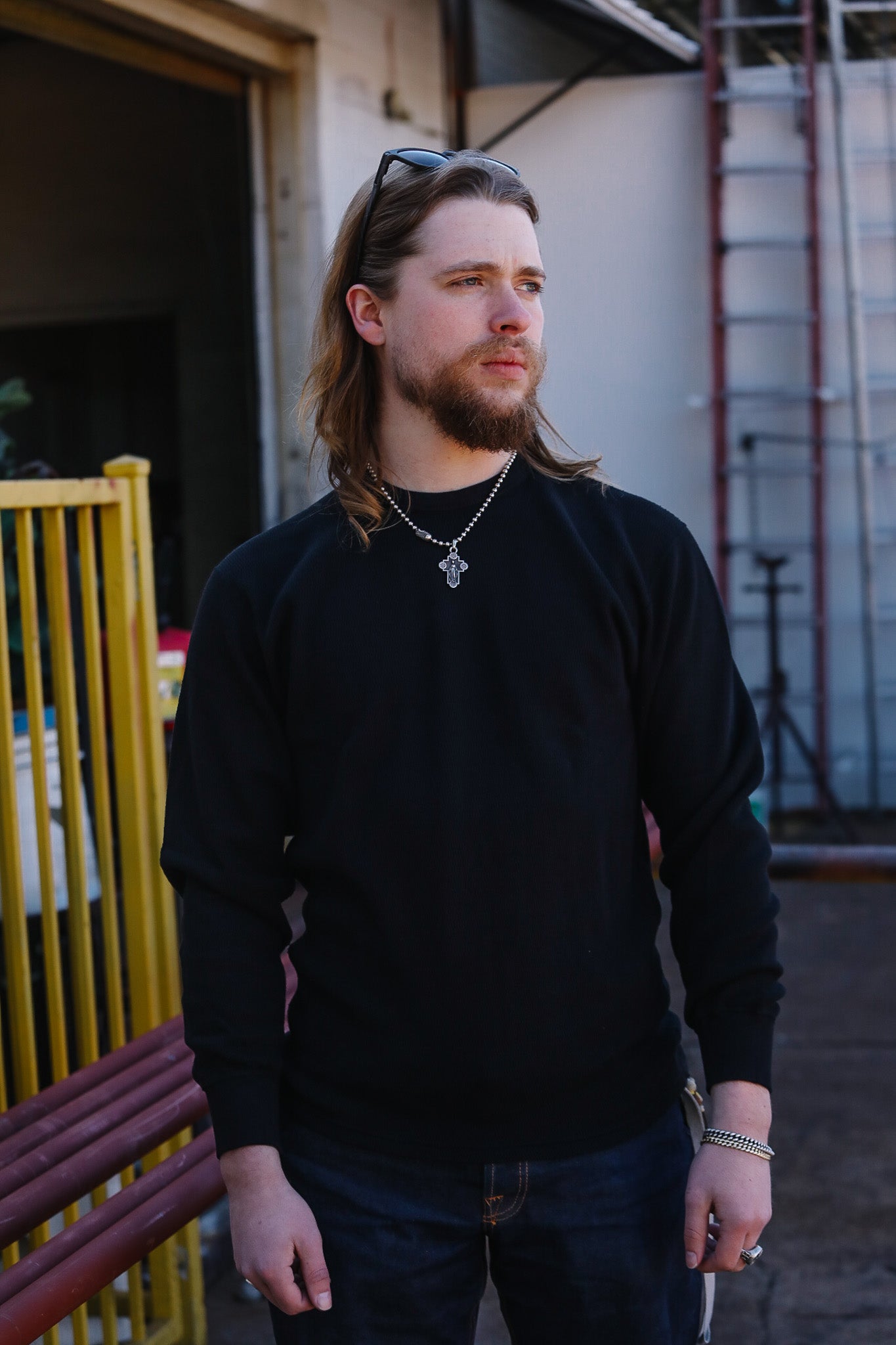 Man wearing a black sweater standing outdoors with a yellow structure and building in the background.