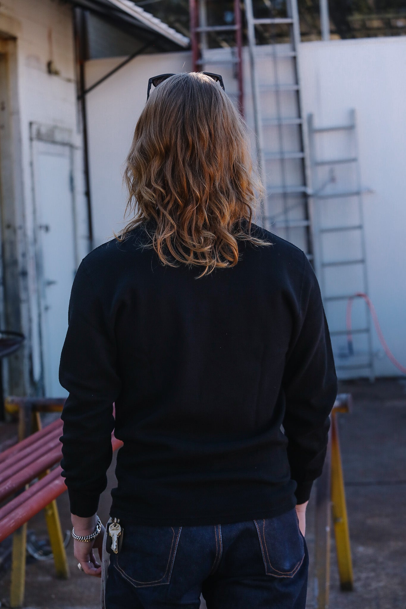 Person wearing a black sweatshirt with long hair, standing in an outdoor setting with a ladder and building in the background.