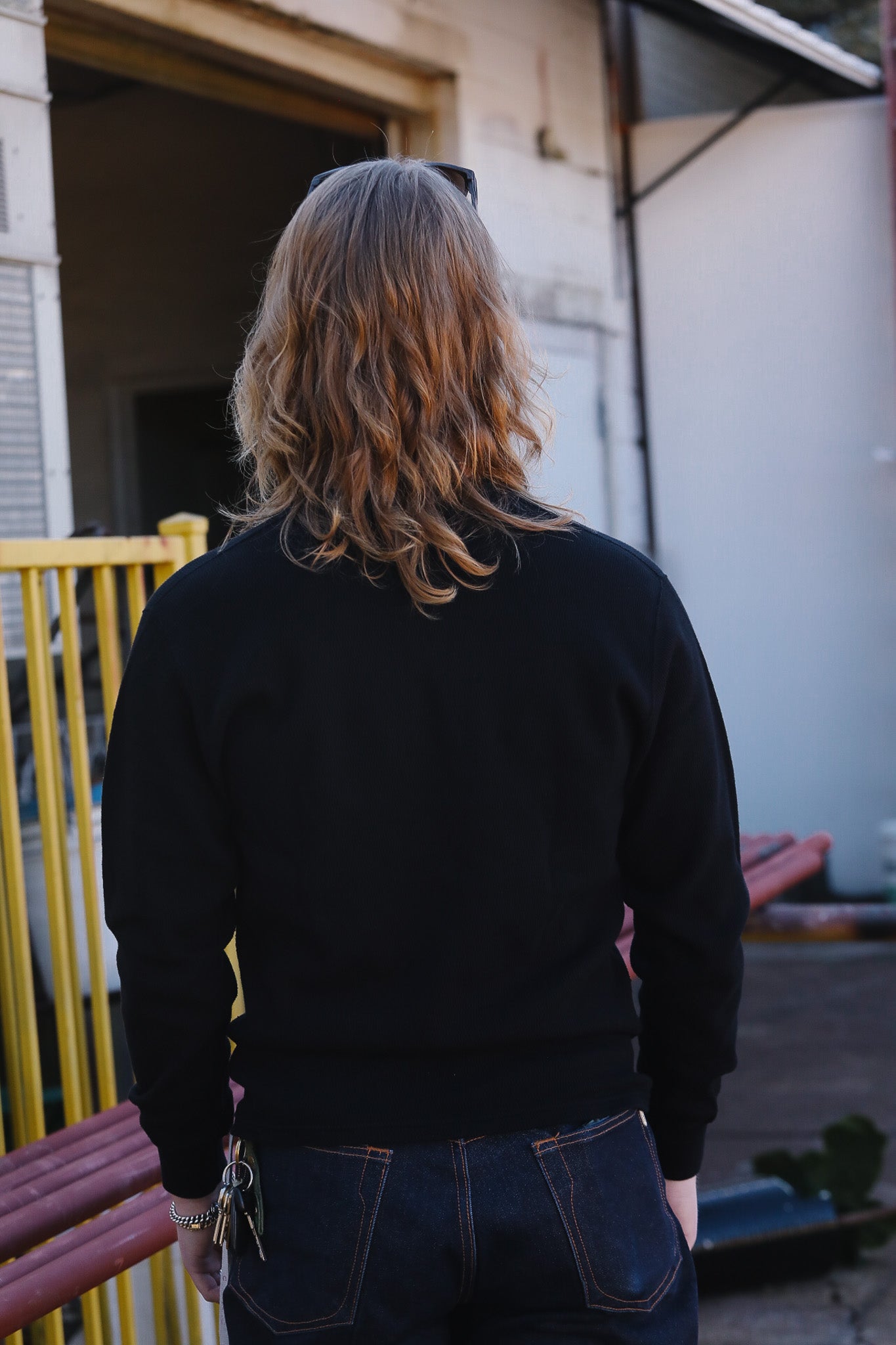 Person wearing a black sweater and blue jeans standing in an outdoor setting with a yellow gate and building in the background.