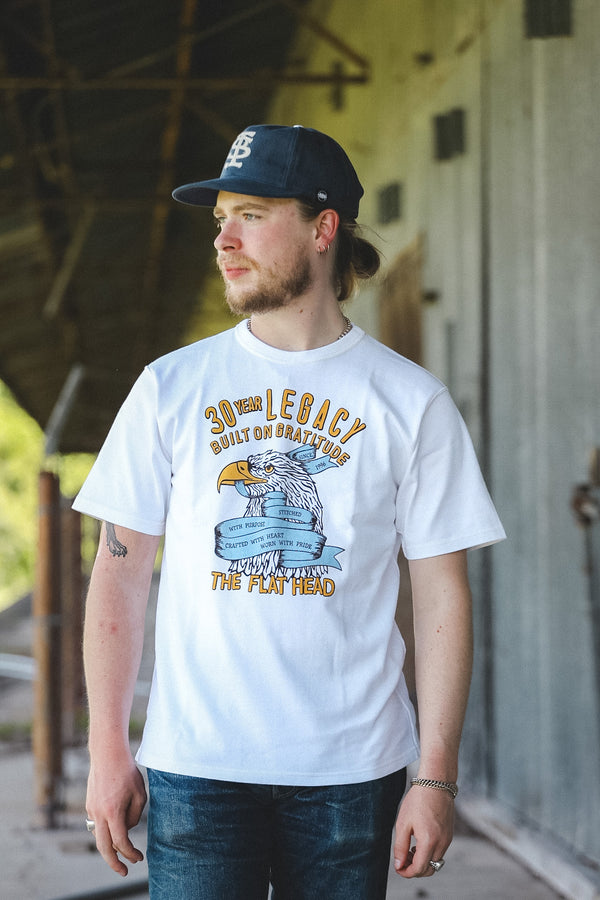 A man wearing a white graphic t-shirt with a blue and yellow eagle design and the text '30th BATTALION ALIASED THE FLAT HERO', paired with a navy blue baseball cap and dark jeans, standing in front of a rustic wooden building.