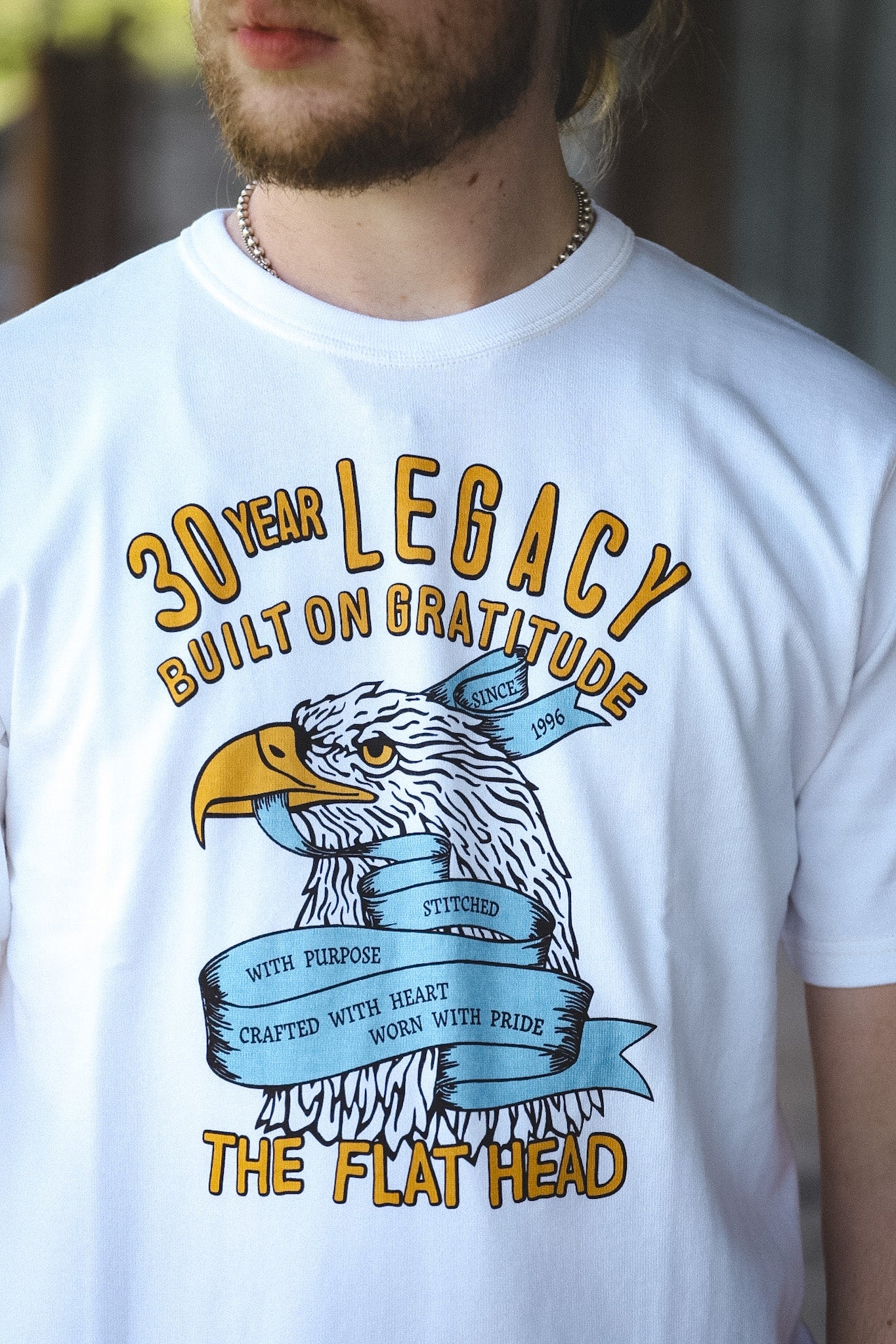 A man wearing a white graphic t-shirt with a yellow and blue eagle design and text reading '30 YEAR LEGACY BUILT ON GRATITUDE' and 'THE FLAT HEAD'.