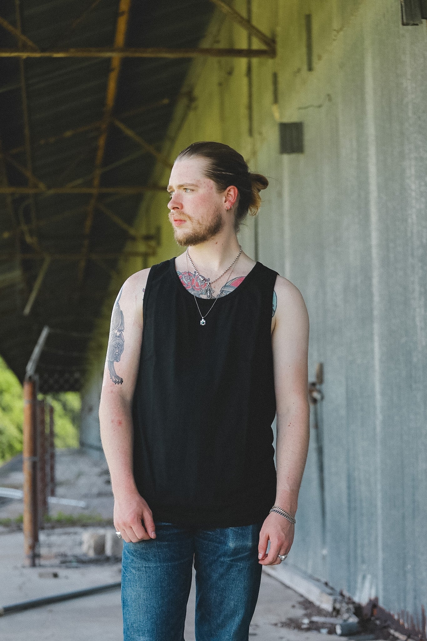 A man with a beard and tattoos wearing a black sleeveless tank top and blue jeans, standing in an industrial covered walkway.