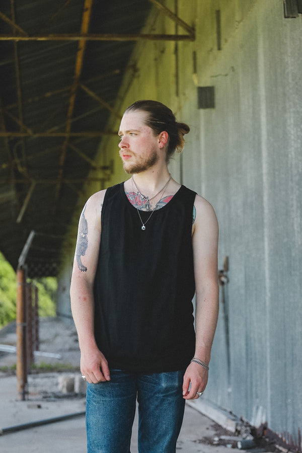 A man with a beard and tattoos wearing a black sleeveless tank top and blue jeans, standing in an industrial covered walkway.