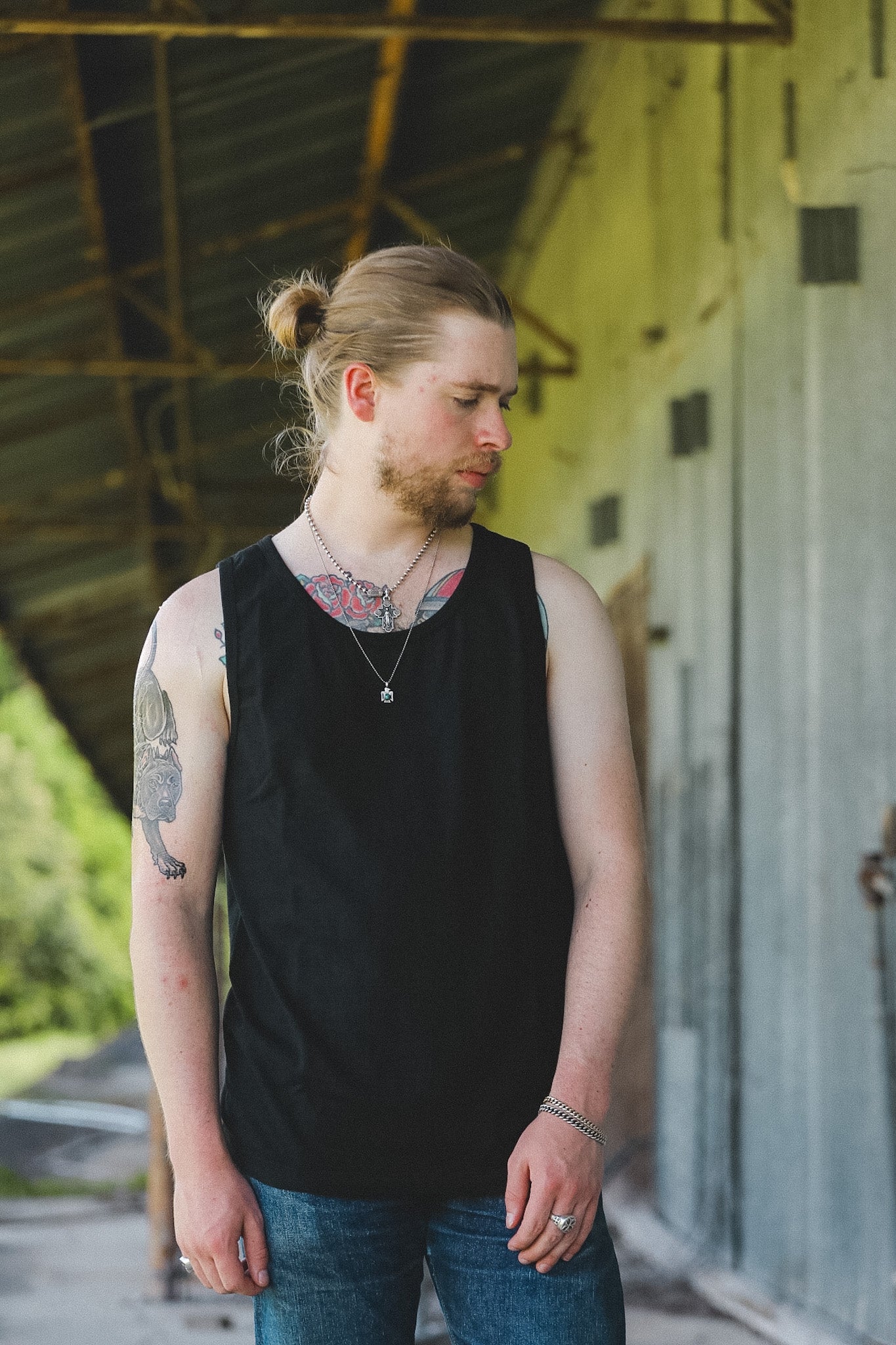 A man with a blonde bun and beard wearing a black sleeveless tank top and blue jeans, standing in an industrial covered walkway.