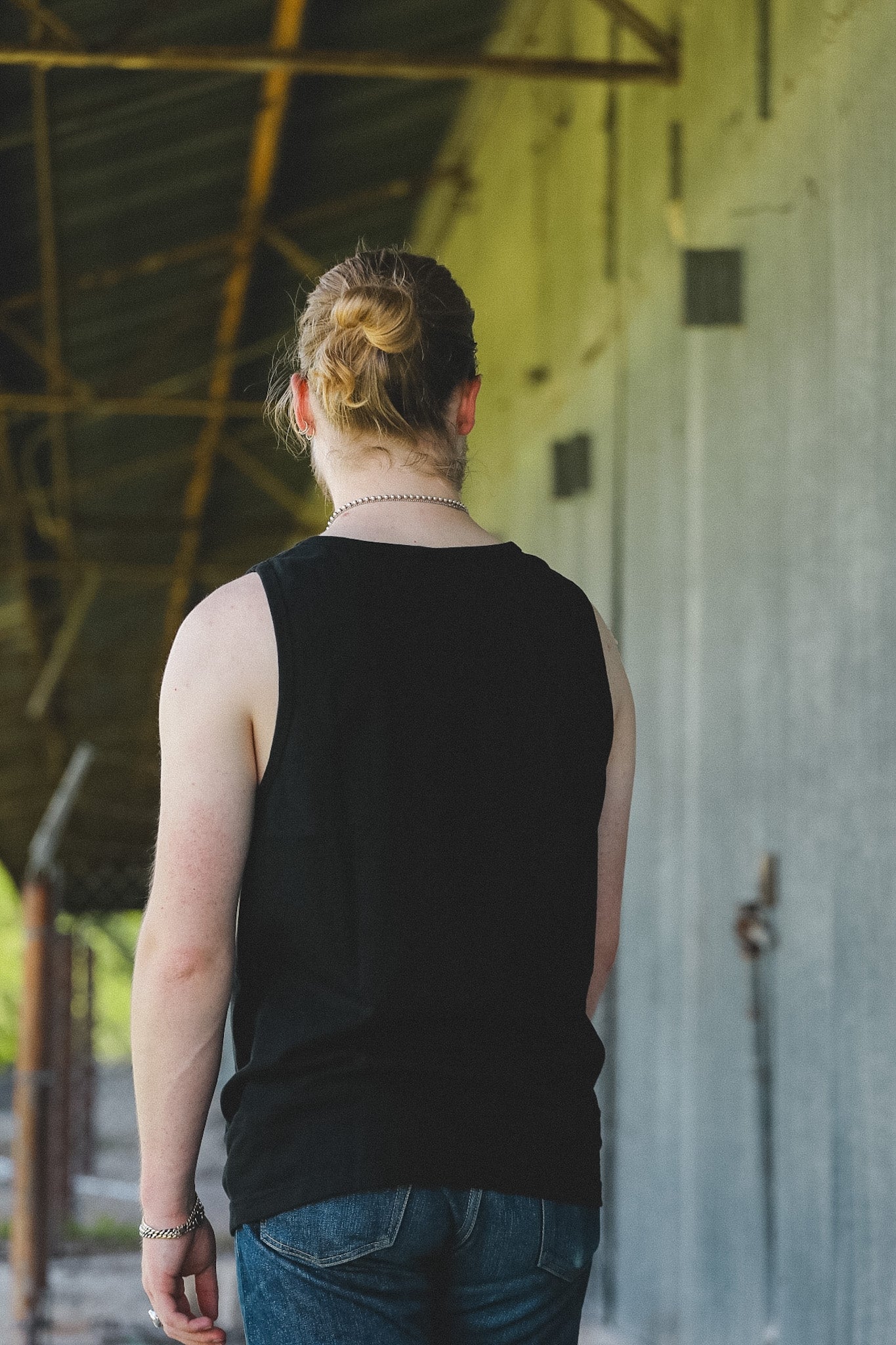 A person viewed from behind wearing a black sleeveless tank top and blue jeans, standing in a covered walkway with a concrete wall on the right.
