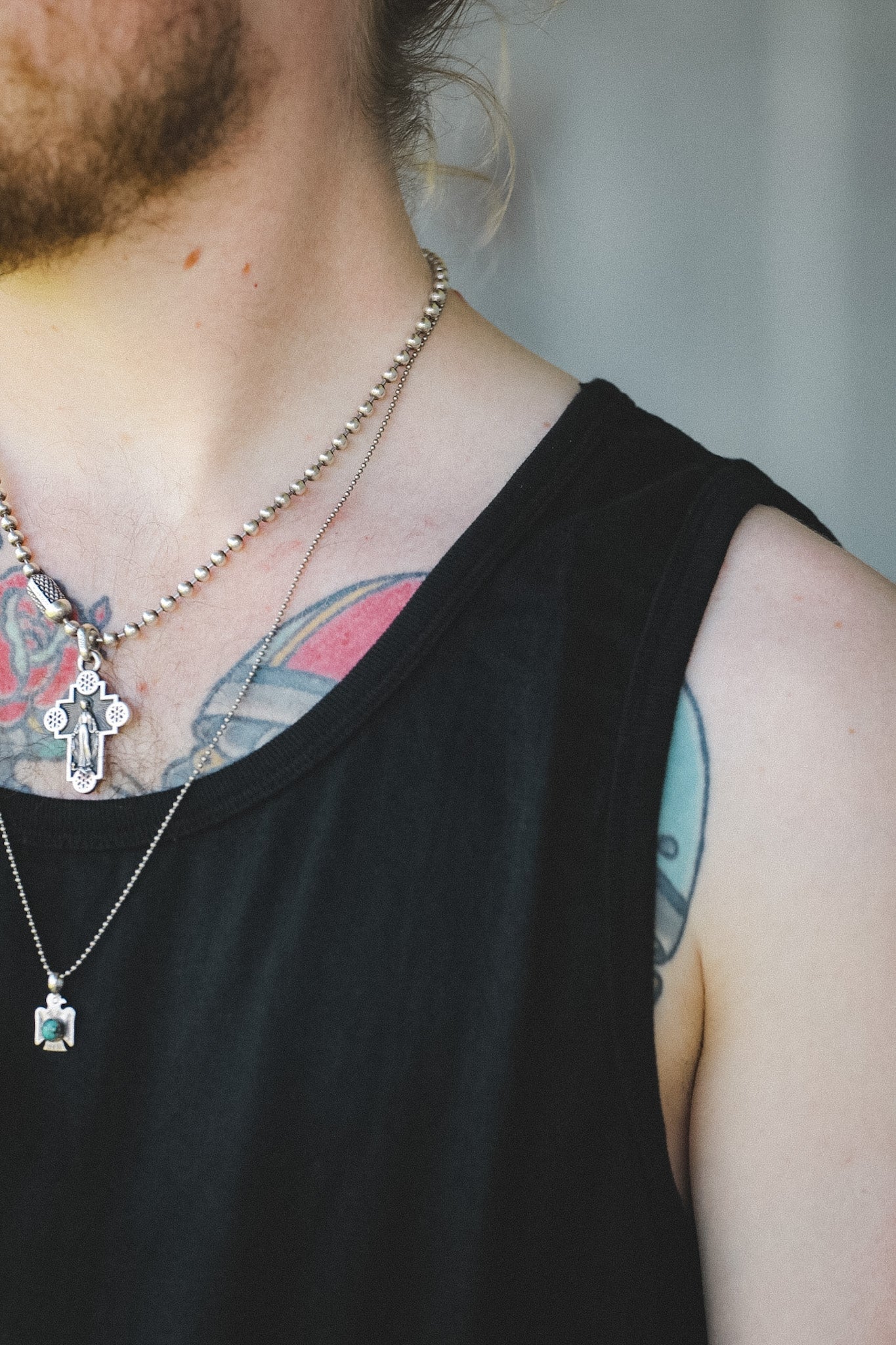 Close-up of a person wearing layered necklaces, including a silver beaded chain with a cross pendant and a silver chain with a small square pendant, over a black tank top.