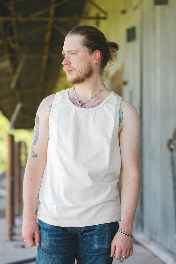 A man with a beard and hair in a ponytail wearing an off-white sleeveless tank top and blue jeans, standing in front of a rustic wooden building.