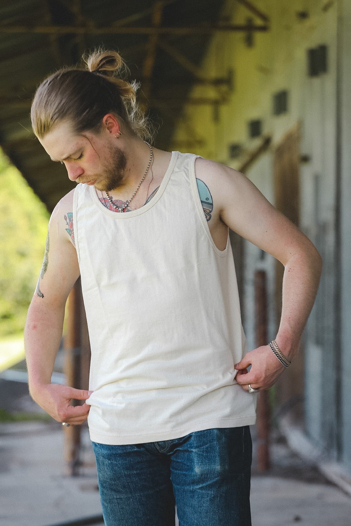 A man with a bun and beard wearing an off-white tank top and blue jeans, standing in front of a rustic building.
