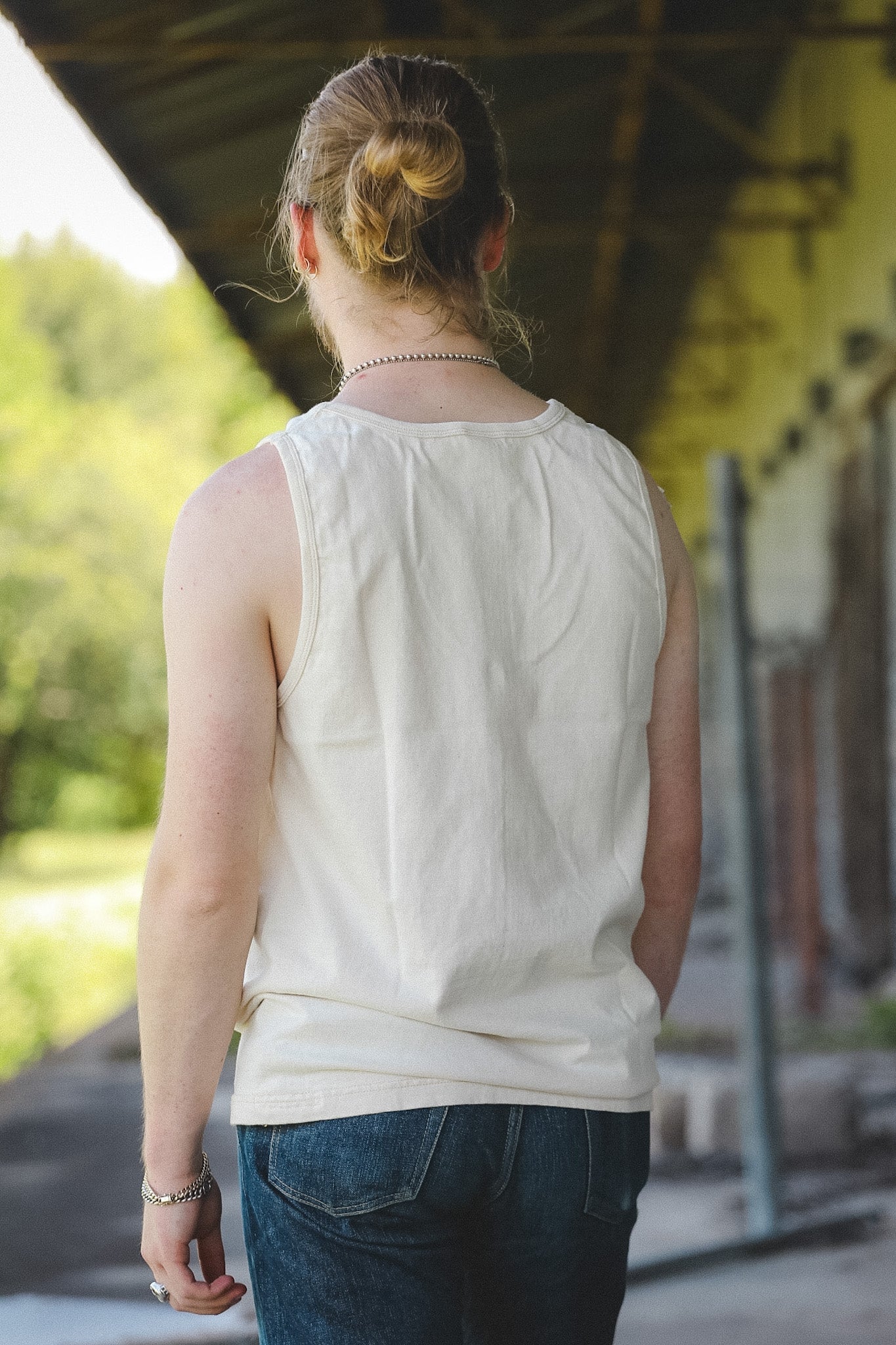 A person viewed from behind wearing a sleeveless cream-colored top and dark blue jeans, standing under a covered walkway.