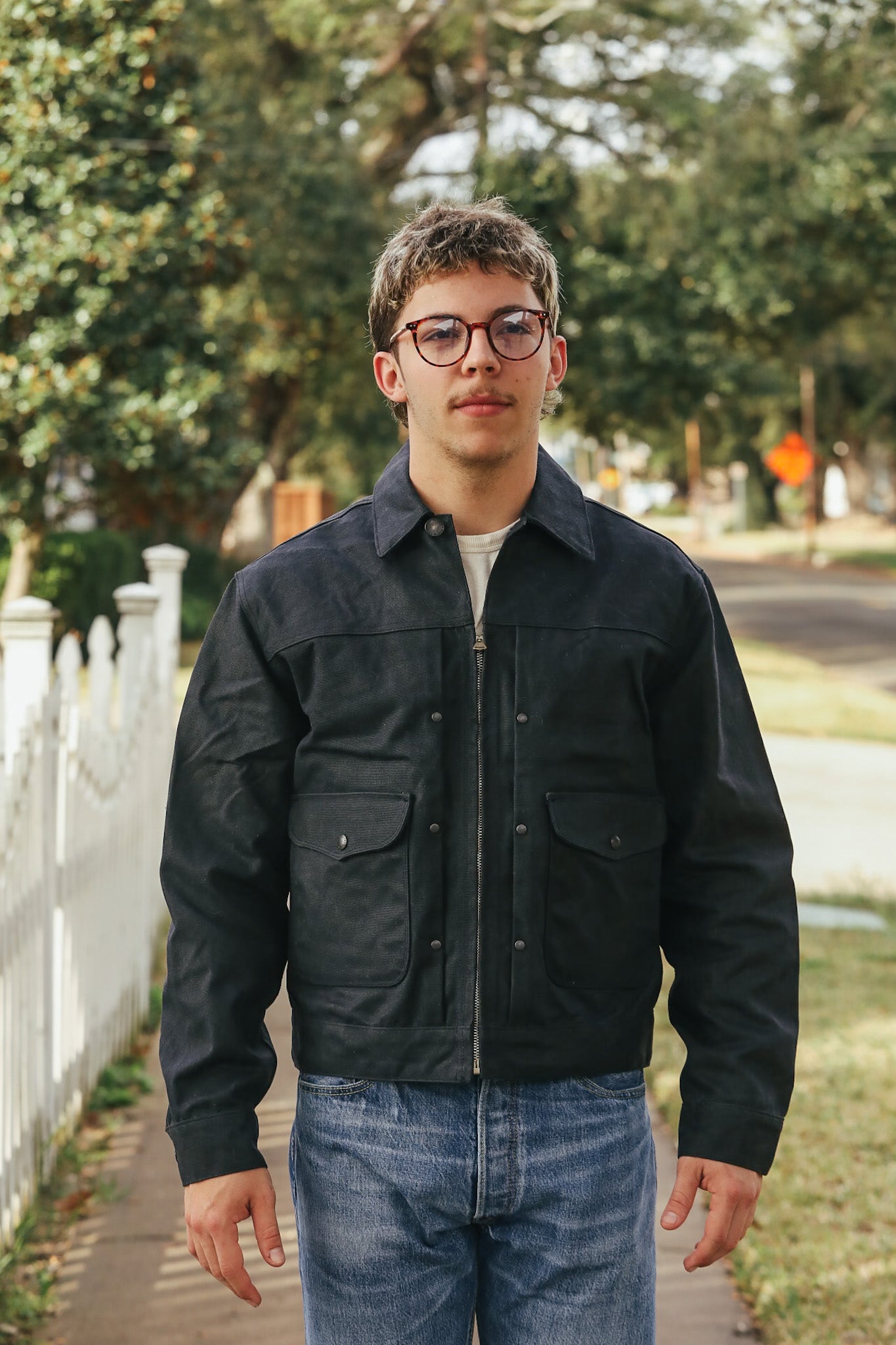 Man wearing a black jacket and glasses standing outdoors with trees and a road in the background