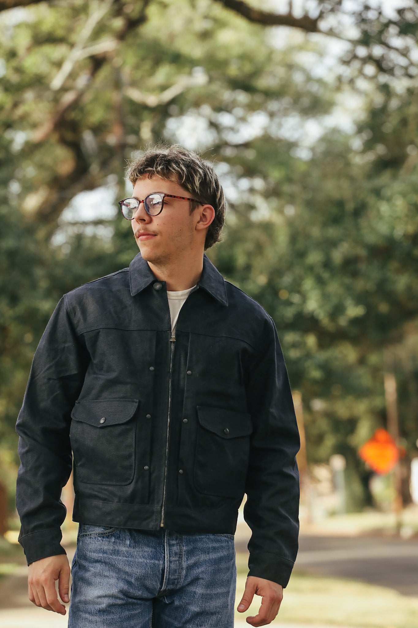 Man wearing a black jacket and glasses standing outdoors with trees in the background