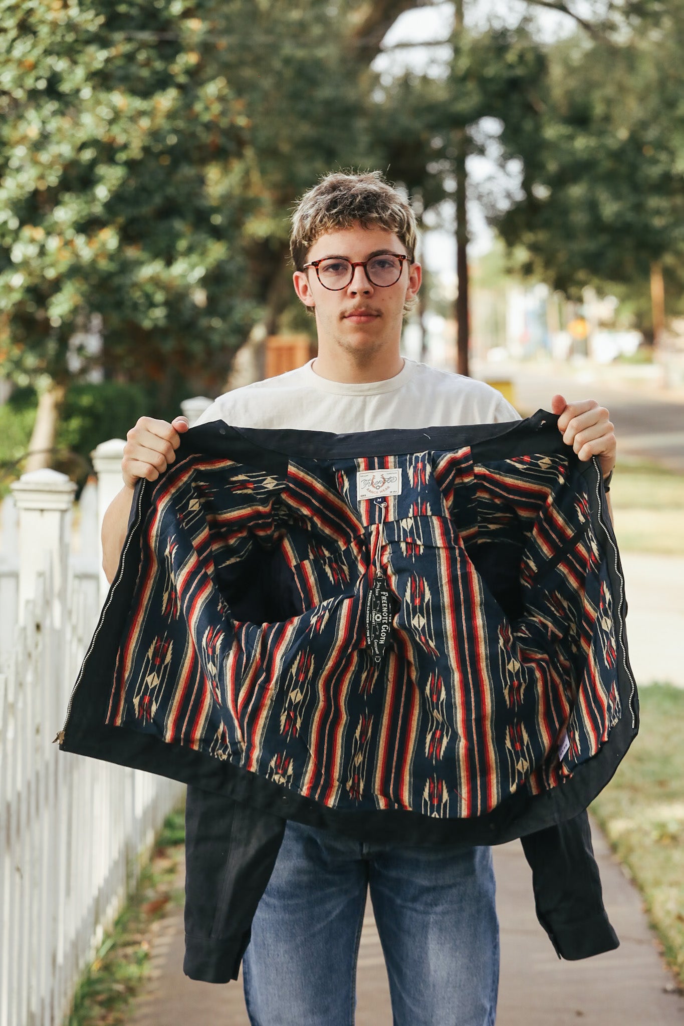 Person holding a patterned jacket outdoors with trees and a fence in the background