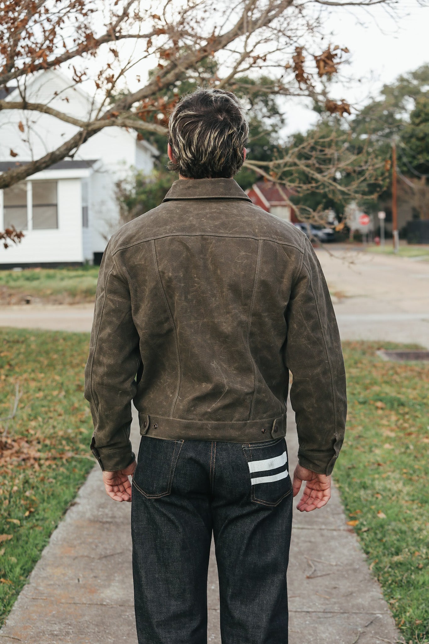 Person wearing a brown jacket and dark pants walking on a sidewalk with trees and houses in the background.