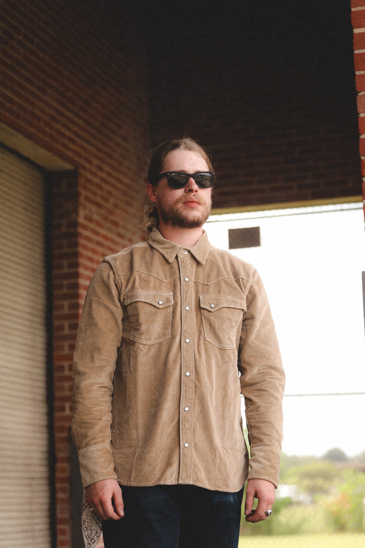 A man wearing a tan corduroy button-down shirt and dark sunglasses, standing in front of a brick building under a covered porch.