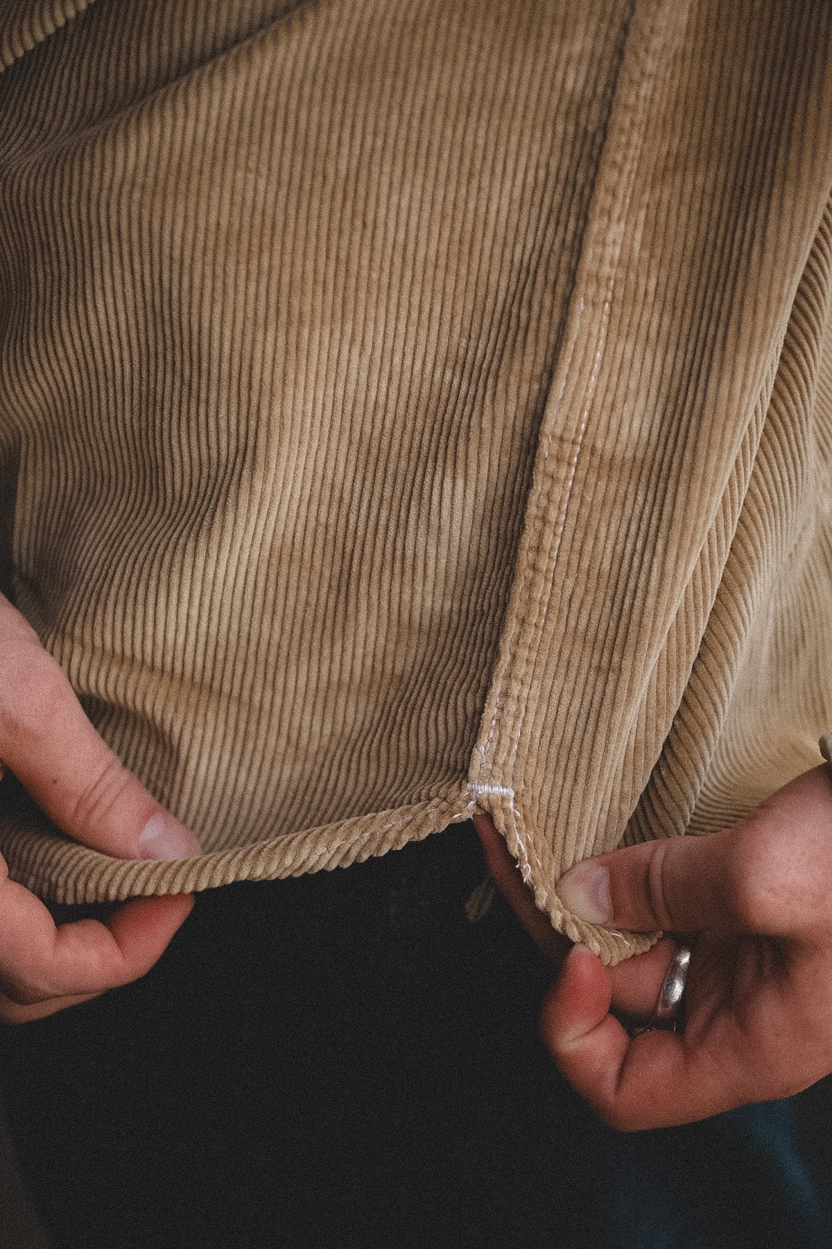 Close-up of a person's hands pulling apart the hem of a tan corduroy jacket to show the fabric's texture and stretch.
