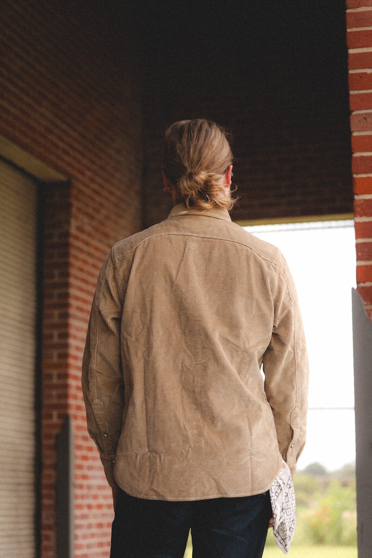 A person viewed from behind wearing a tan corduroy button-up shirt and dark pants, standing in a brick doorway.